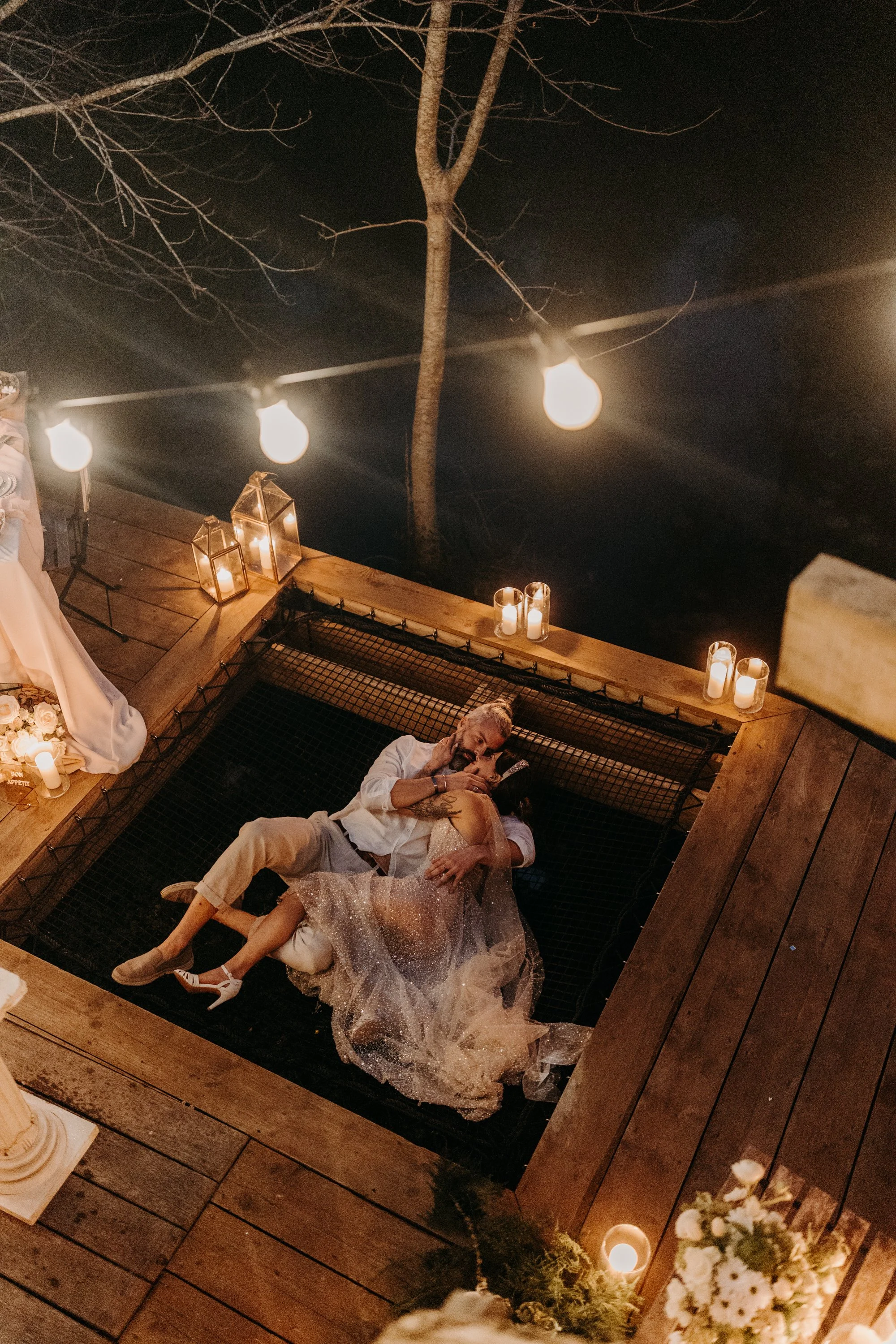Un couple danse et s'embrasse sur une plateforme en plein air décorée de bougies et de lanternes, sous des guirlandes lumineuses la nuit.