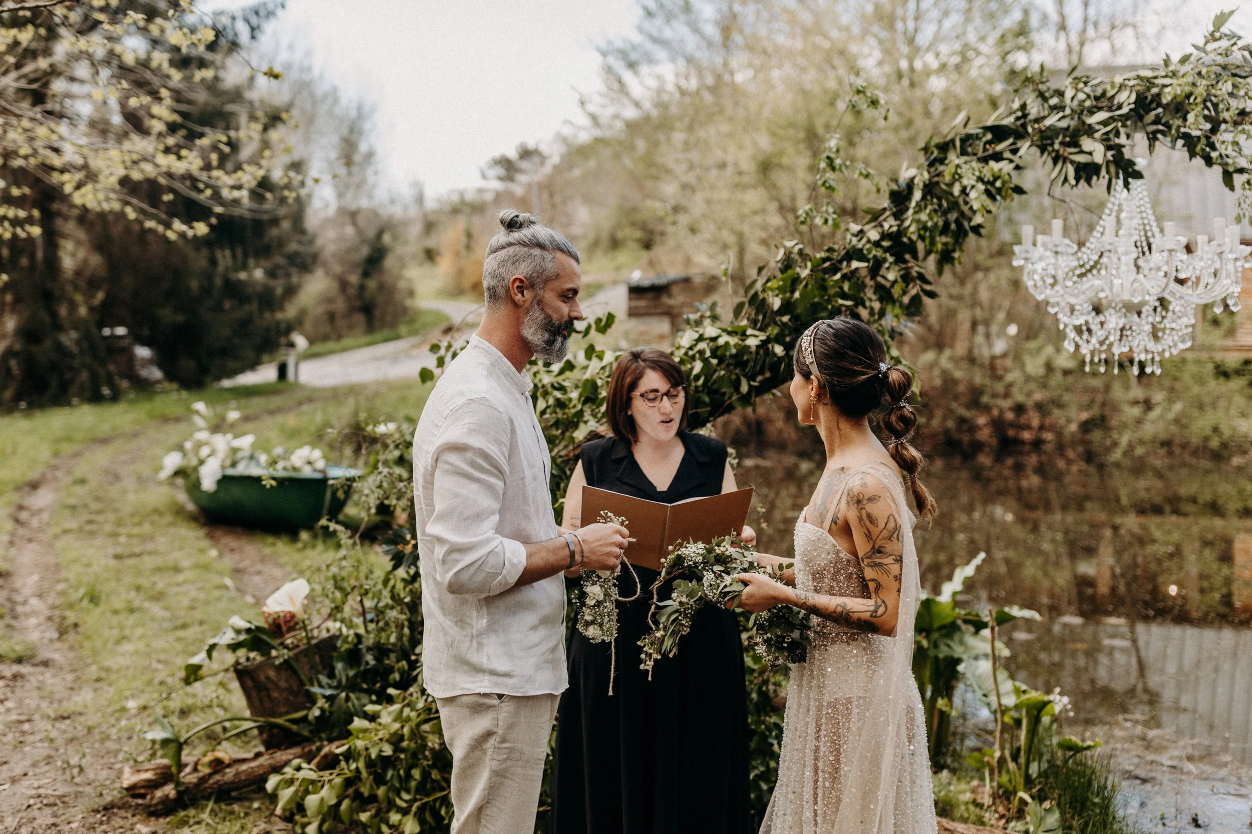 Un couple de mariés lors de leur cérémonie de mariage en plein air, avec un officiant, près d'un étang et décoré de fleurs et d'un chandelier en cristal.