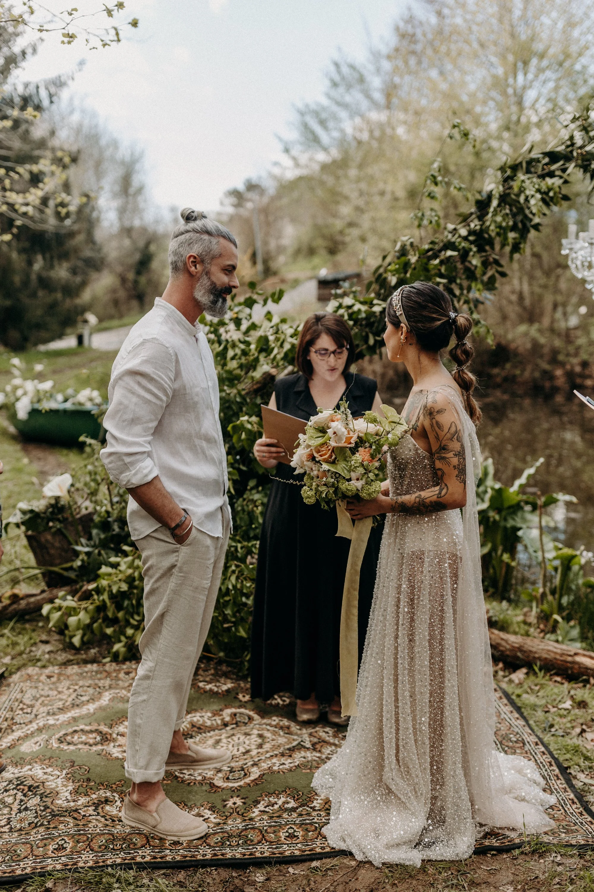 Un couple échangeant leurs vœux de mariage lors d'une cérémonie en plein air, avec une officiant, dans un décor naturel avec des arbres et des fleurs.