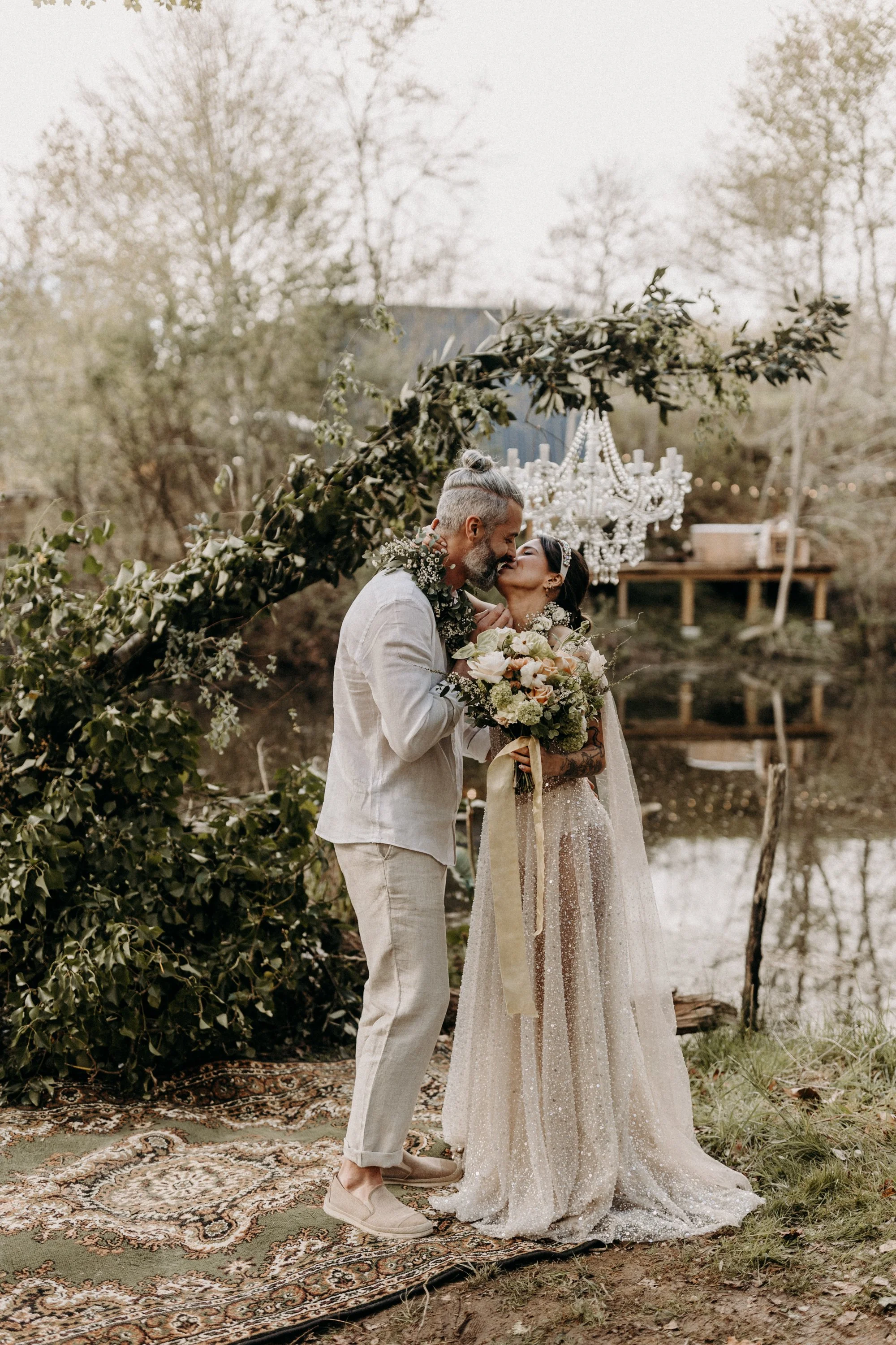 Un couple de mariés échangeant un baiser en plein air, avec un décor naturel et une chandelier suspendu.