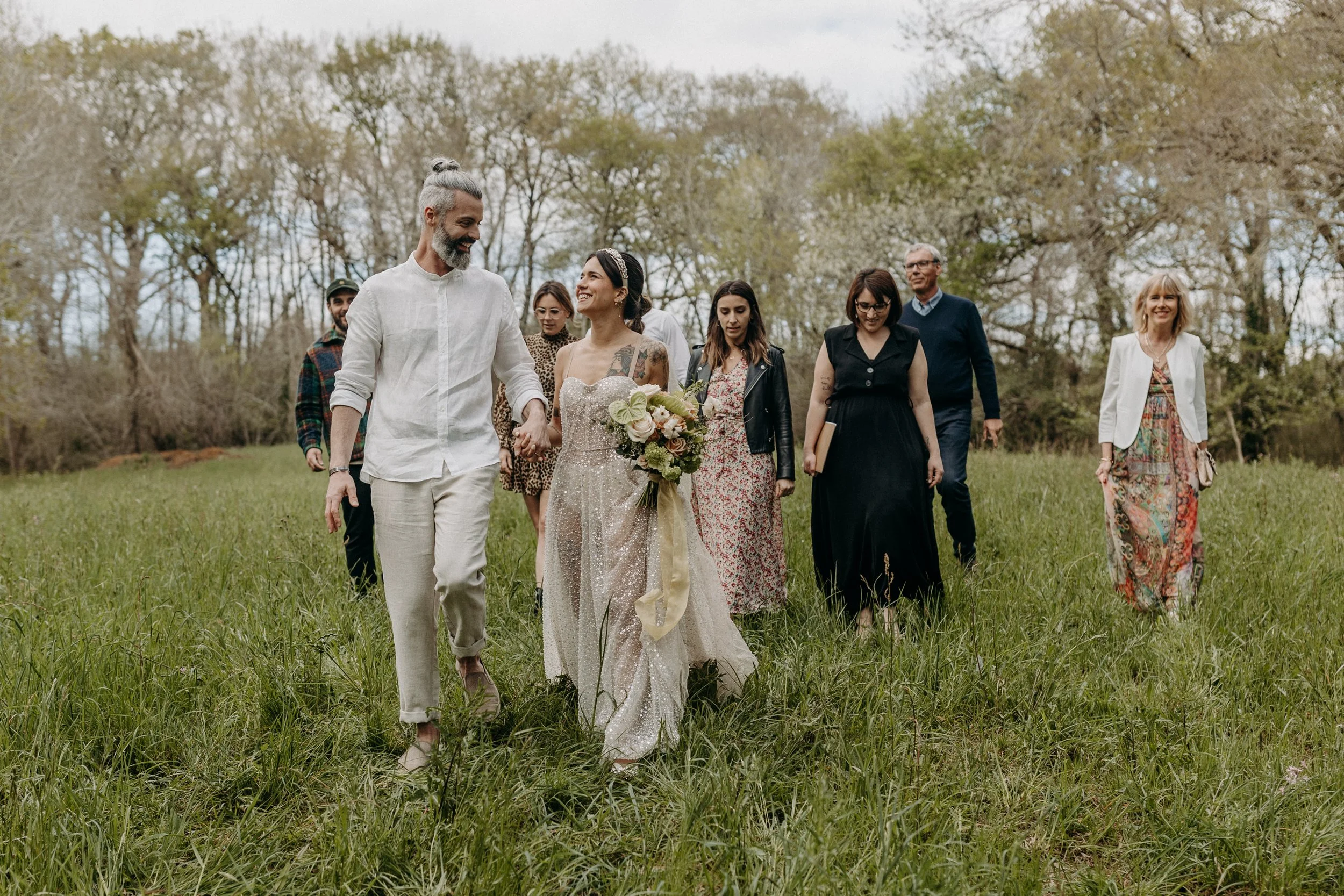 Un groupe de personnes marchant dans un champ lors d'une célébration, avec une femme habillée en robe de mariée tenant un bouquet de fleurs, entourée d'amis et famille dans un cadre naturel.