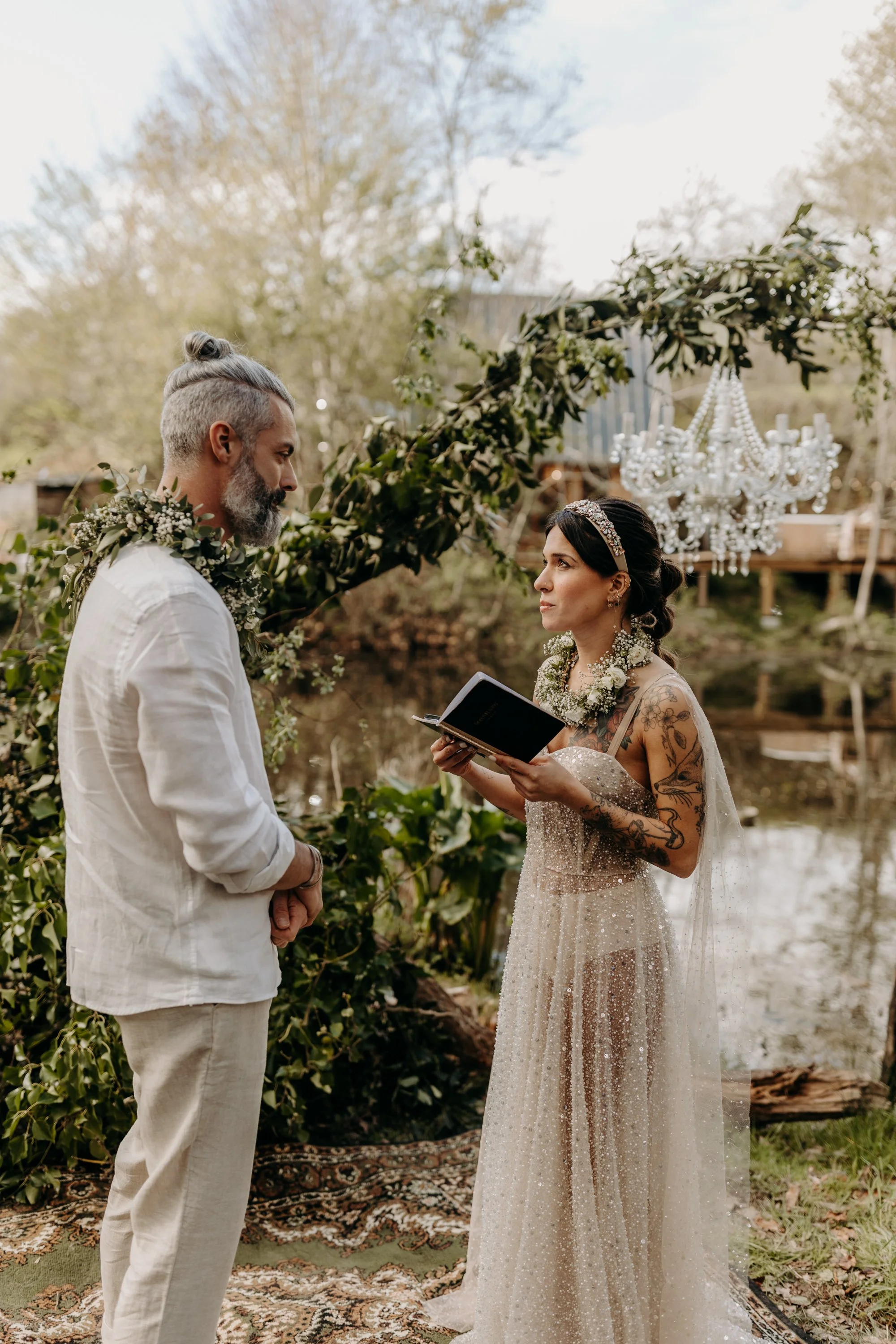 Un couple lors d'une cérémonie de mariage en extérieur, avec la femme tenant un livre, sous une arche décorée de verdure, avec un lustre suspendu en arrière-plan.