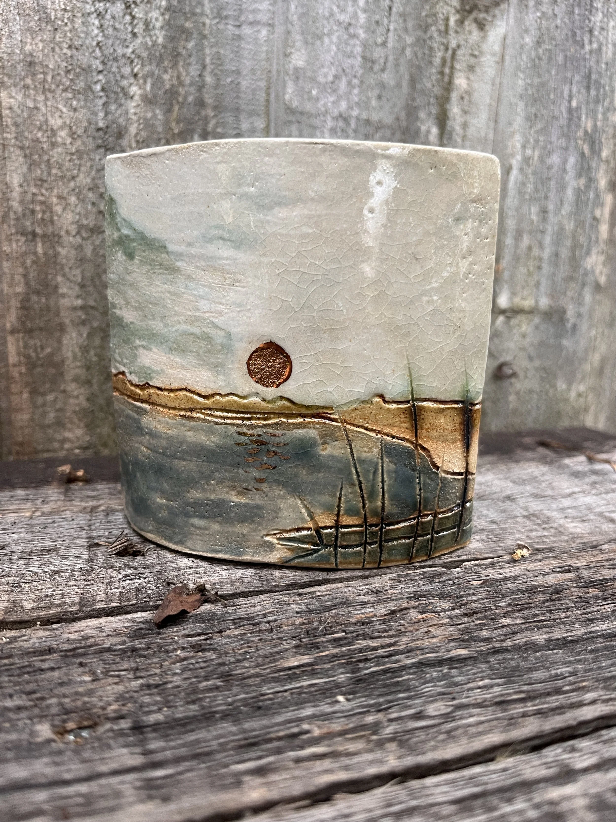 A ceramic cup with landscape artwork featuring a setting or rising sun, water, and grass, placed on weathered wood with a wooden fence in the background.