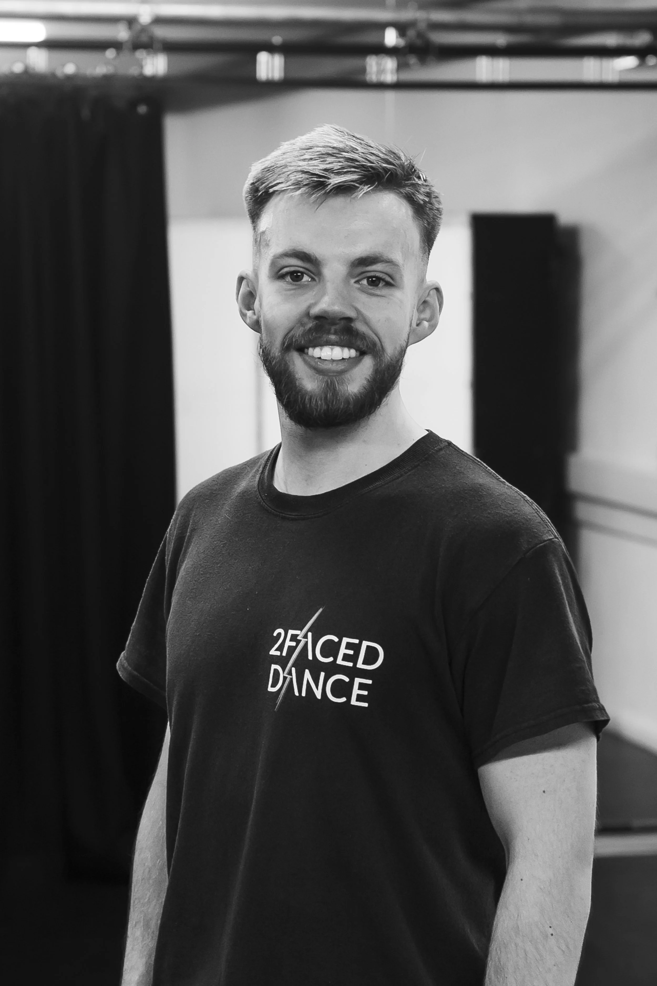 A young man with a beard and short hair smiling at the camera, wearing a black t-shirt with the words '2 Faced Dance' printed on it, standing indoors with a blurred background.