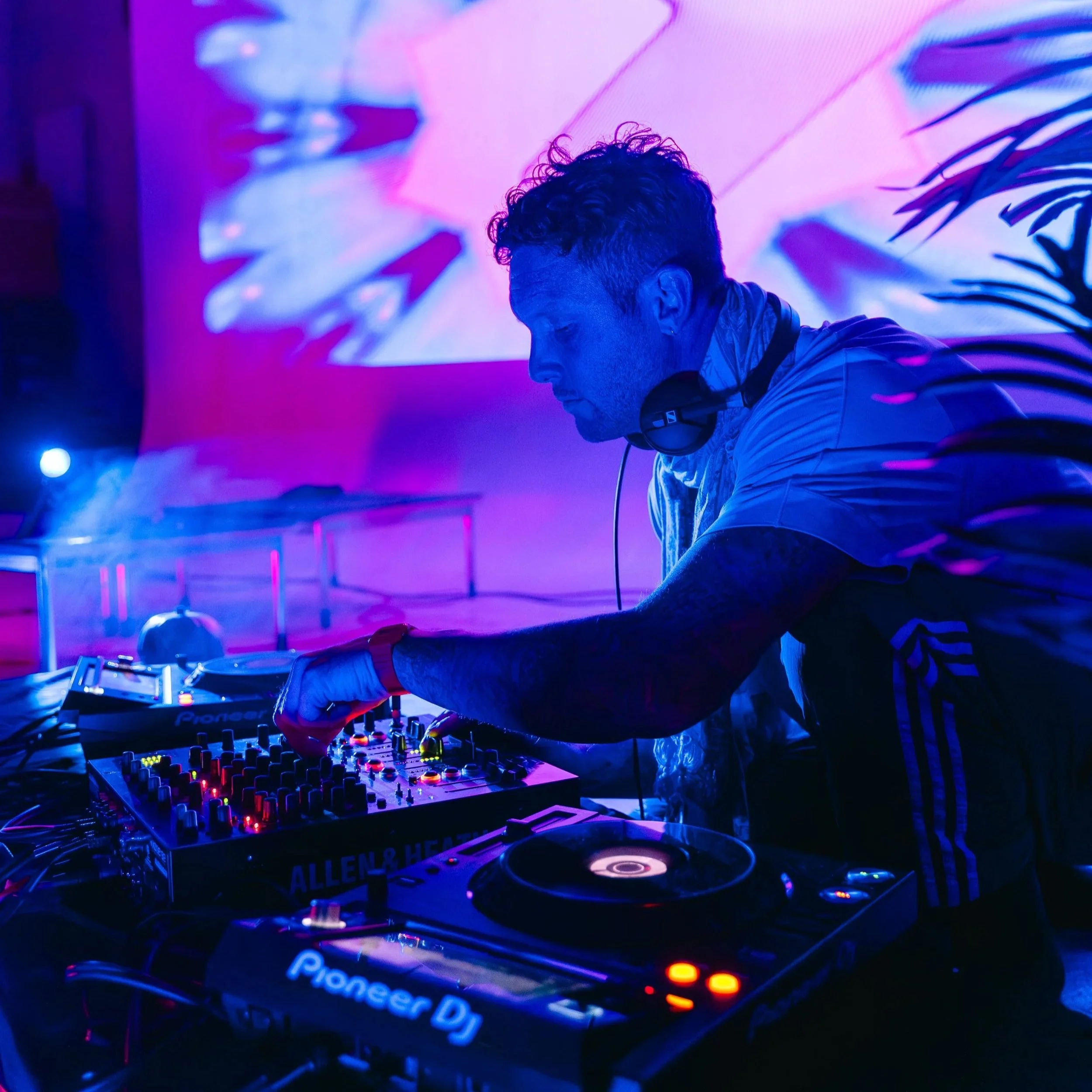 A DJ with curly hair wearing headphones, focused on a Pioneer DJ mixer in a dimly lit setting with vibrant purple and pink lighting, a large digital screen in the background, and a plant silhouette on the right.