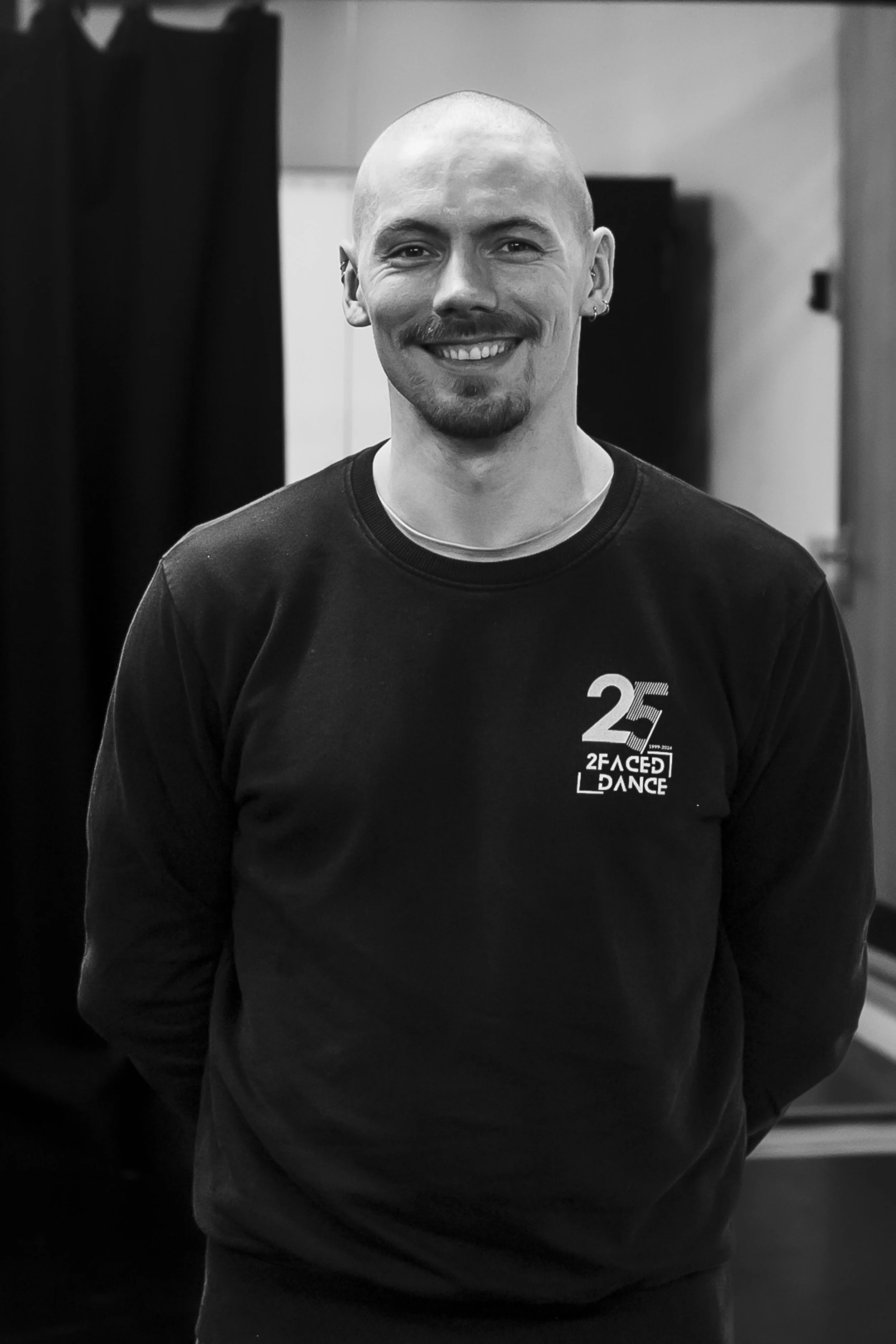A smiling man with a shaved head, goatee, and earrings, wearing a sweatshirt with a '25' logo and text '2Faced Dance' on it, standing indoors.