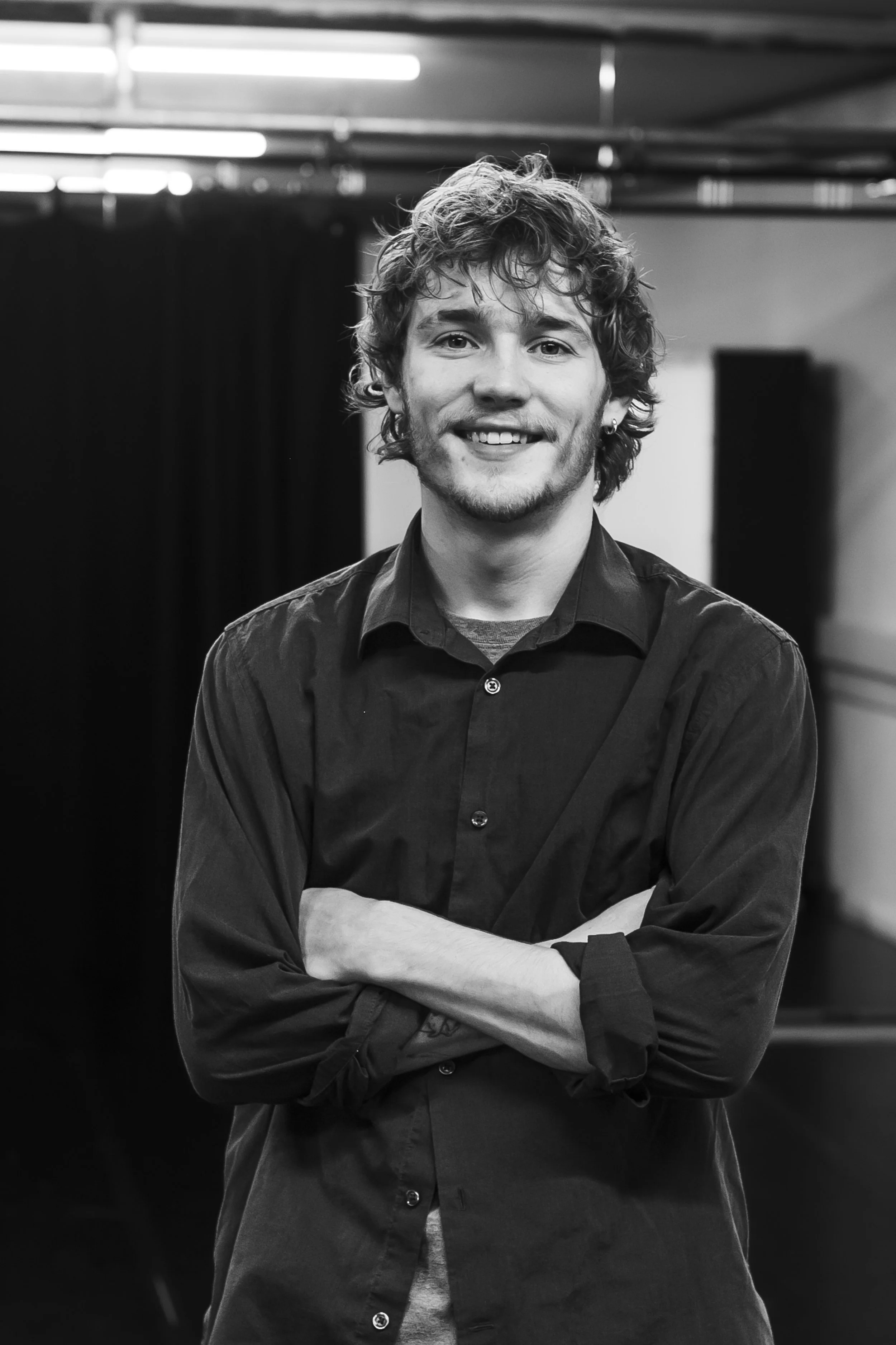 A smiling young man with curly hair and a beard, standing with his arms crossed in a black shirt in a room with dark curtains and some light fixtures.