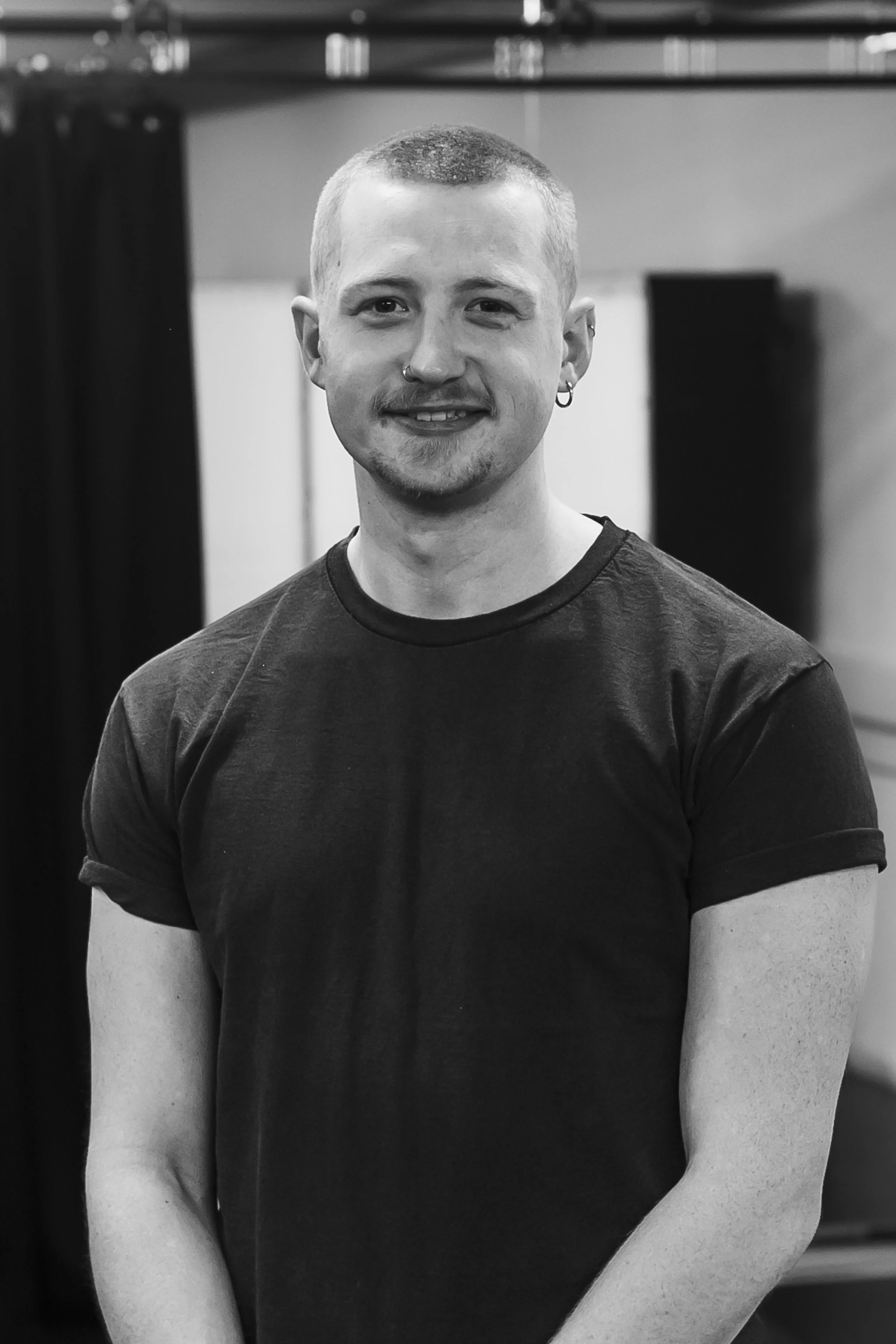A young man with short hair, a small beard, and earrings, smiling while standing indoors in front of a background with blurred shelves or equipment.