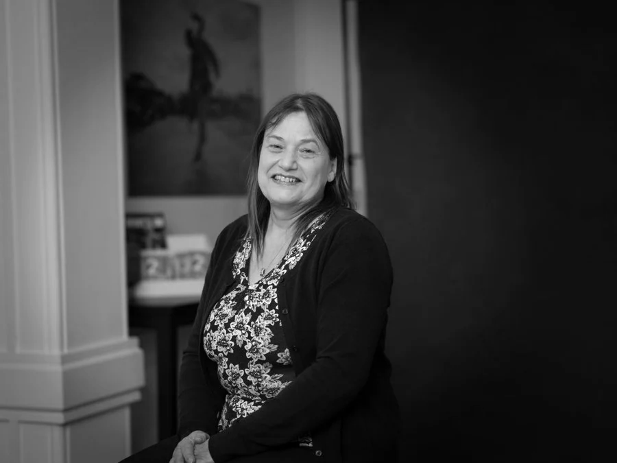 A woman with shoulder-length hair sitting and smiling, wearing a floral top and dark cardigan, with a painting and a table in the background.