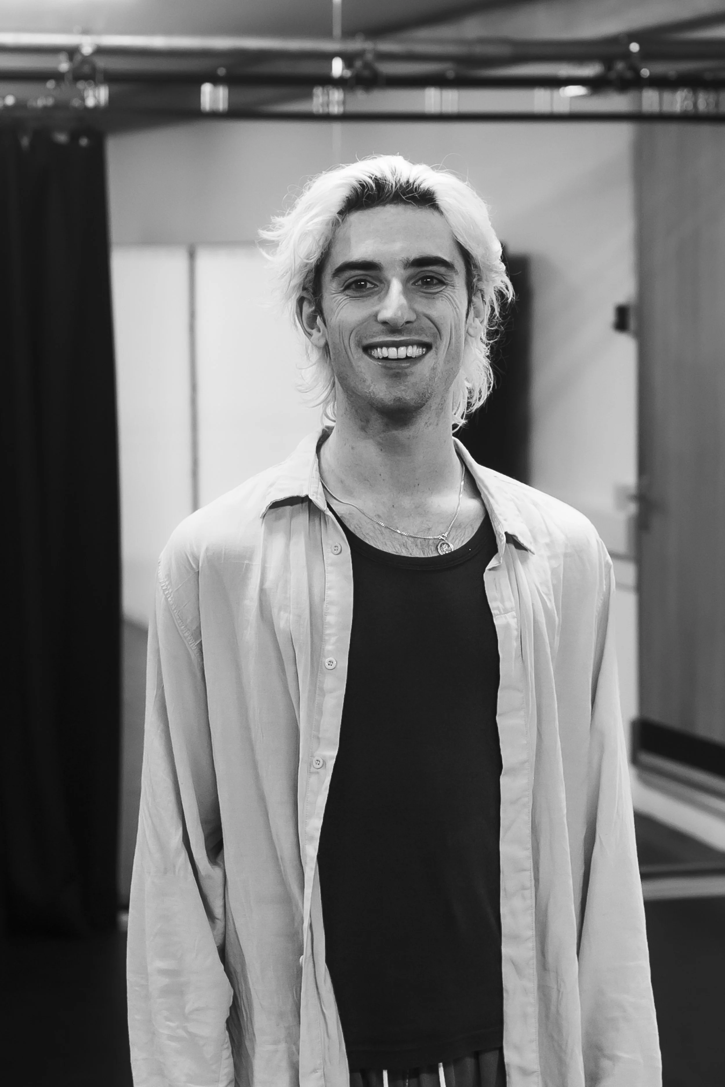 A young man with long, wavy, platinum blonde hair smiling at the camera in an indoor setting.