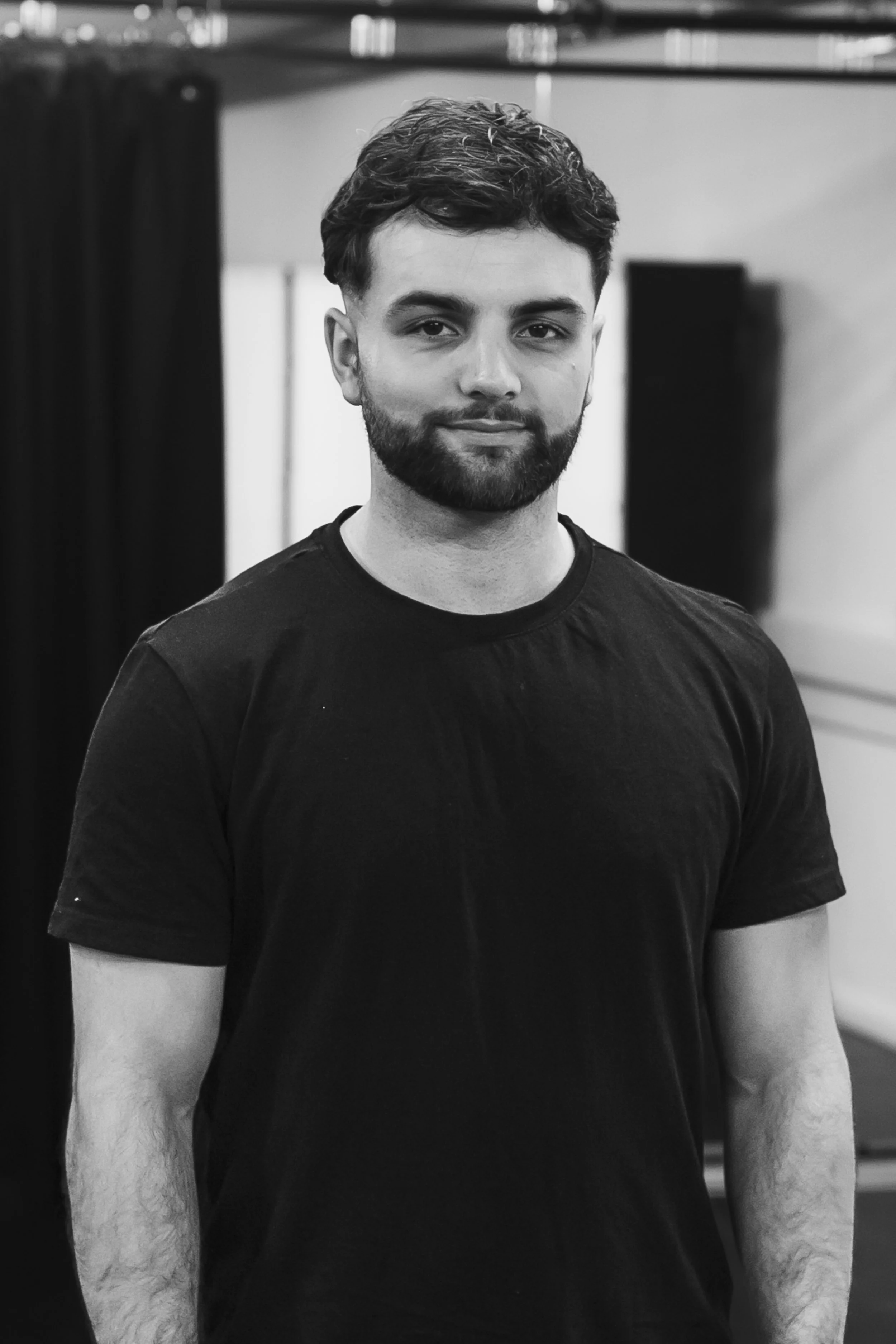 Black and white portrait of a young man with a beard, wearing a black t-shirt, standing indoors.