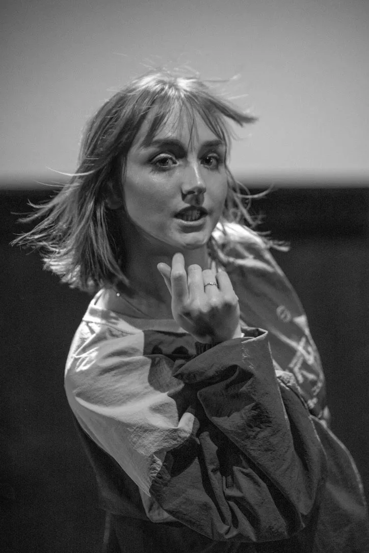 Black and white photo of a woman with shoulder-length hair, wearing a jacket, looking at the camera with a hand near her face, in a dramatic pose.