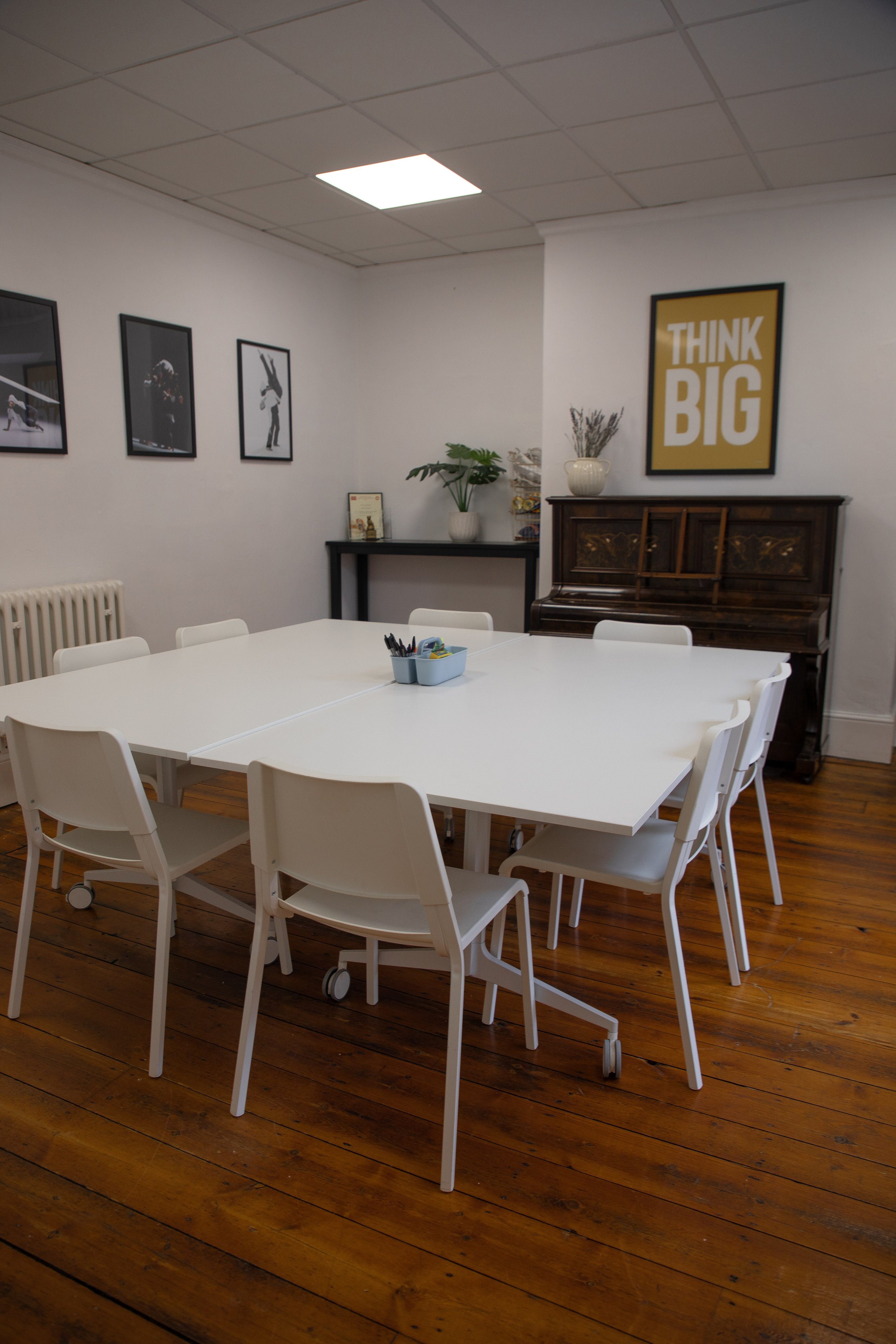A meeting room with white tables and chairs, a hardwood floor, white walls decorated with framed photos, a piano, a large yellow and white poster reading 'THINK BIG', and some potted plants.