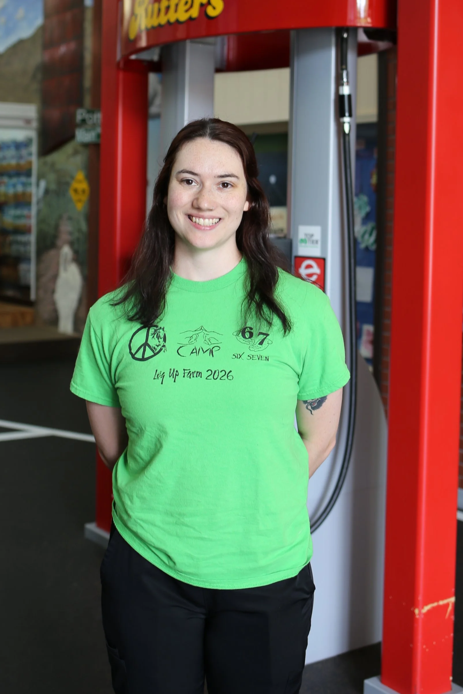 A smiling woman with long dark hair, wearing a bright green T-shirt with text and graphics and black pants, standing in front of a Redbox DVD rental kiosk.