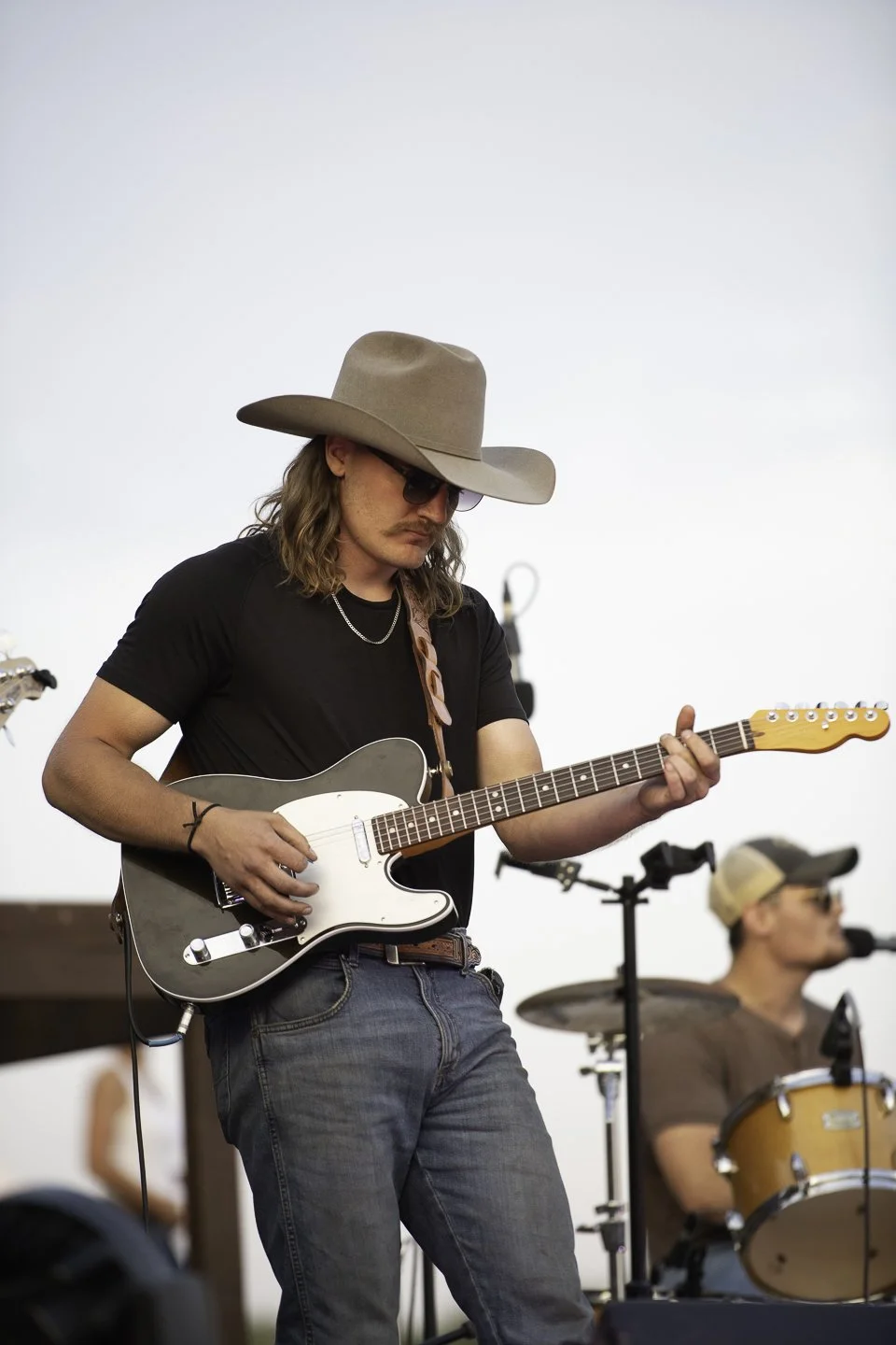 A musician wearing a large beige cowboy hat, sunglasses, a black t-shirt, and jeans playing an electric guitar during a live outdoor performance.