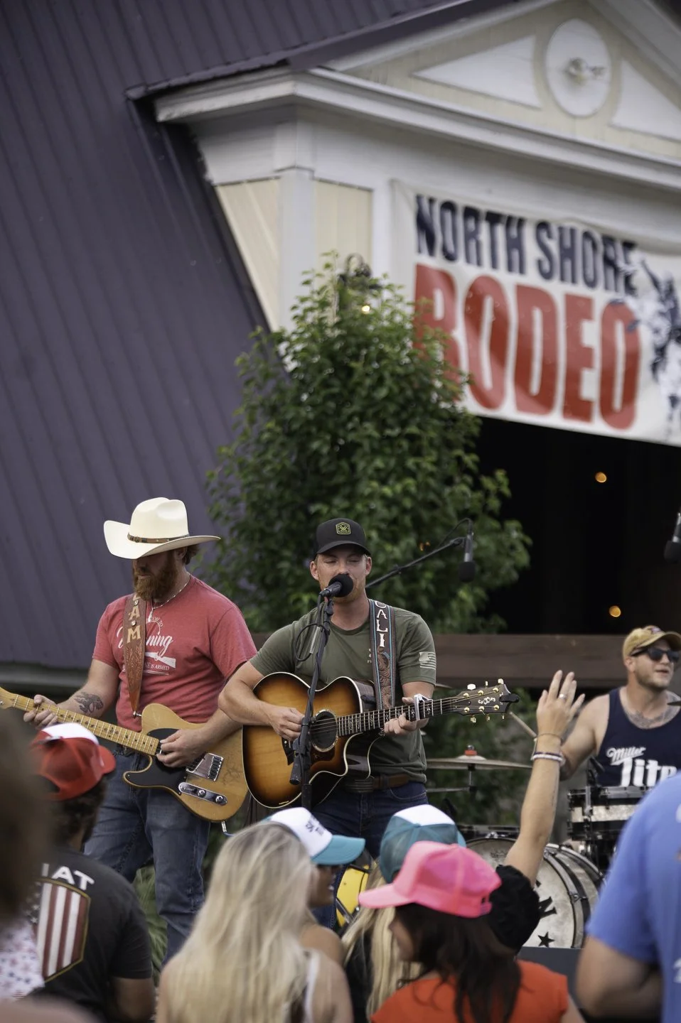 A live country music performance at North Shore Rodeo featuring three male musicians on stage with an audience watching, some wearing colorful caps.
