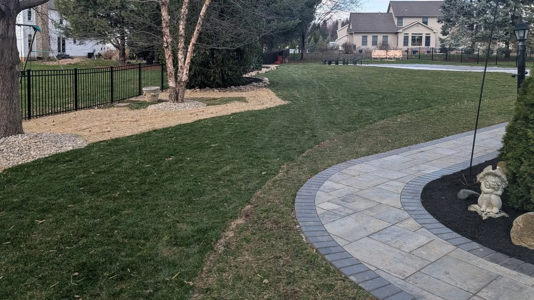 Backyard with lawn, trees, a curved stone patio, a decorative statue of a dog, and a black metal fence. In the background are houses, trees, and a cloudy sky.