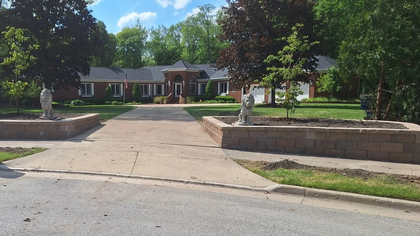 A large brick house with a driveway leading to the entrance, flanked by two raised flower beds with statues of lions, trees, and a blue sky with clouds.