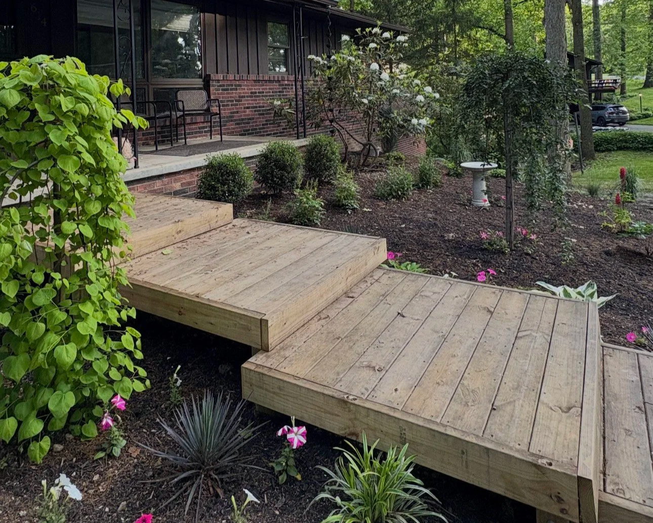 Newly built wooden deck with steps leading to a porch of a house surrounded by a landscaped garden with flowering plants, bushes, and trees.