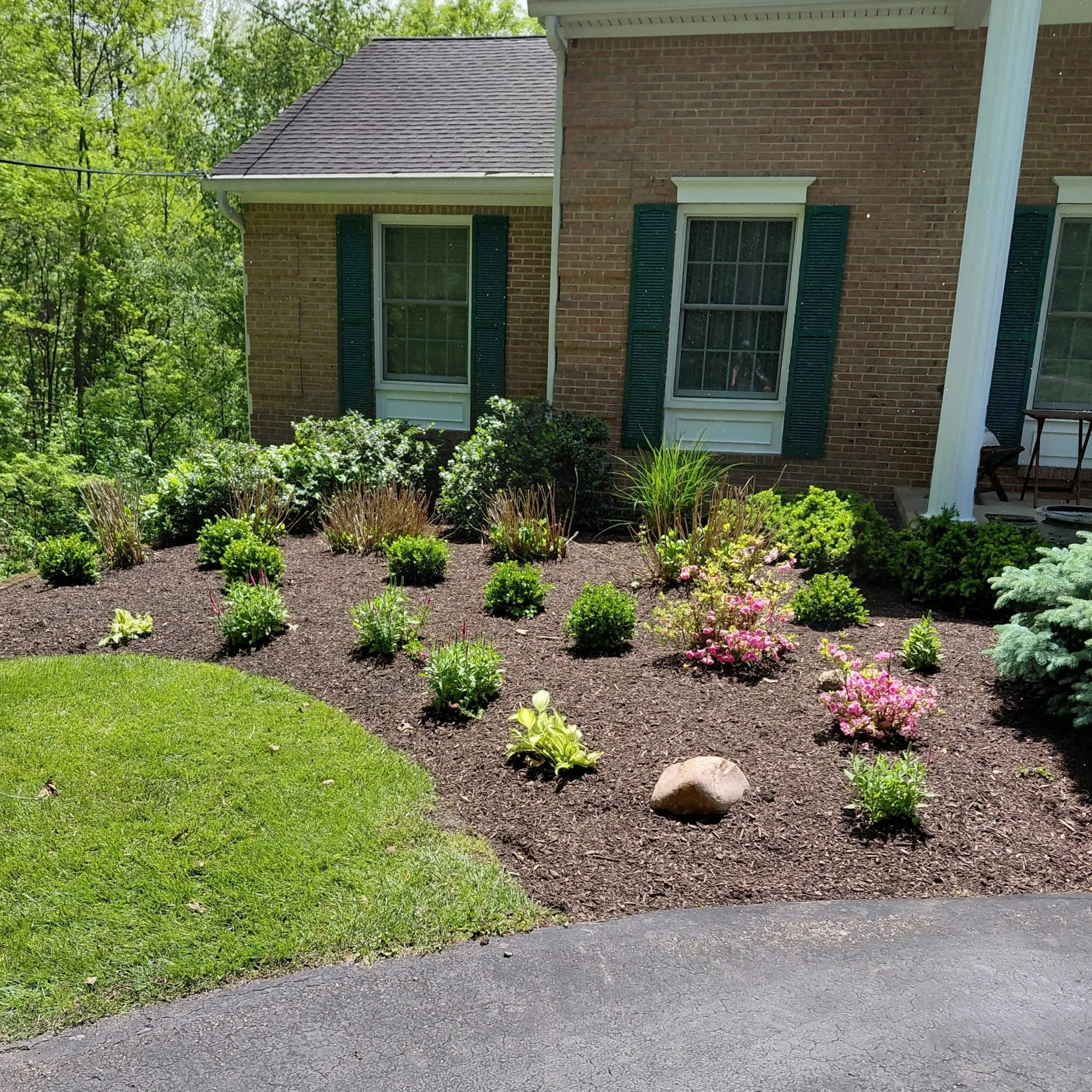 View of a brick house with two windows and dark green shutters, surrounded by a landscaped garden with various plants, bushes, and pink flowers, with lush green trees in the background and part of a paved driveway or street in the foreground.