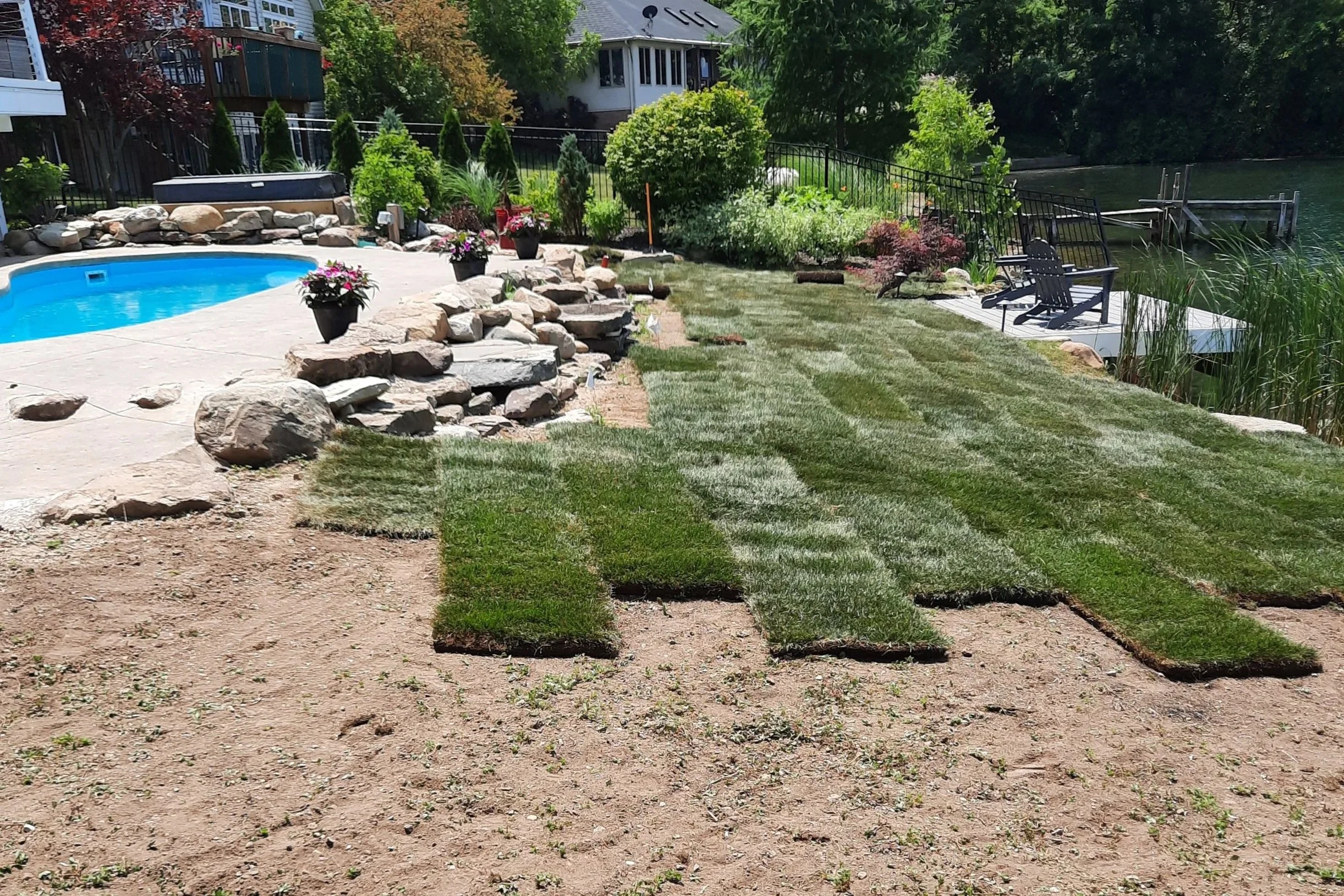 Backyard with a swimming pool, rock border, potted plants, a lawn, a wooden dock by a lake, and surrounding trees and houses.