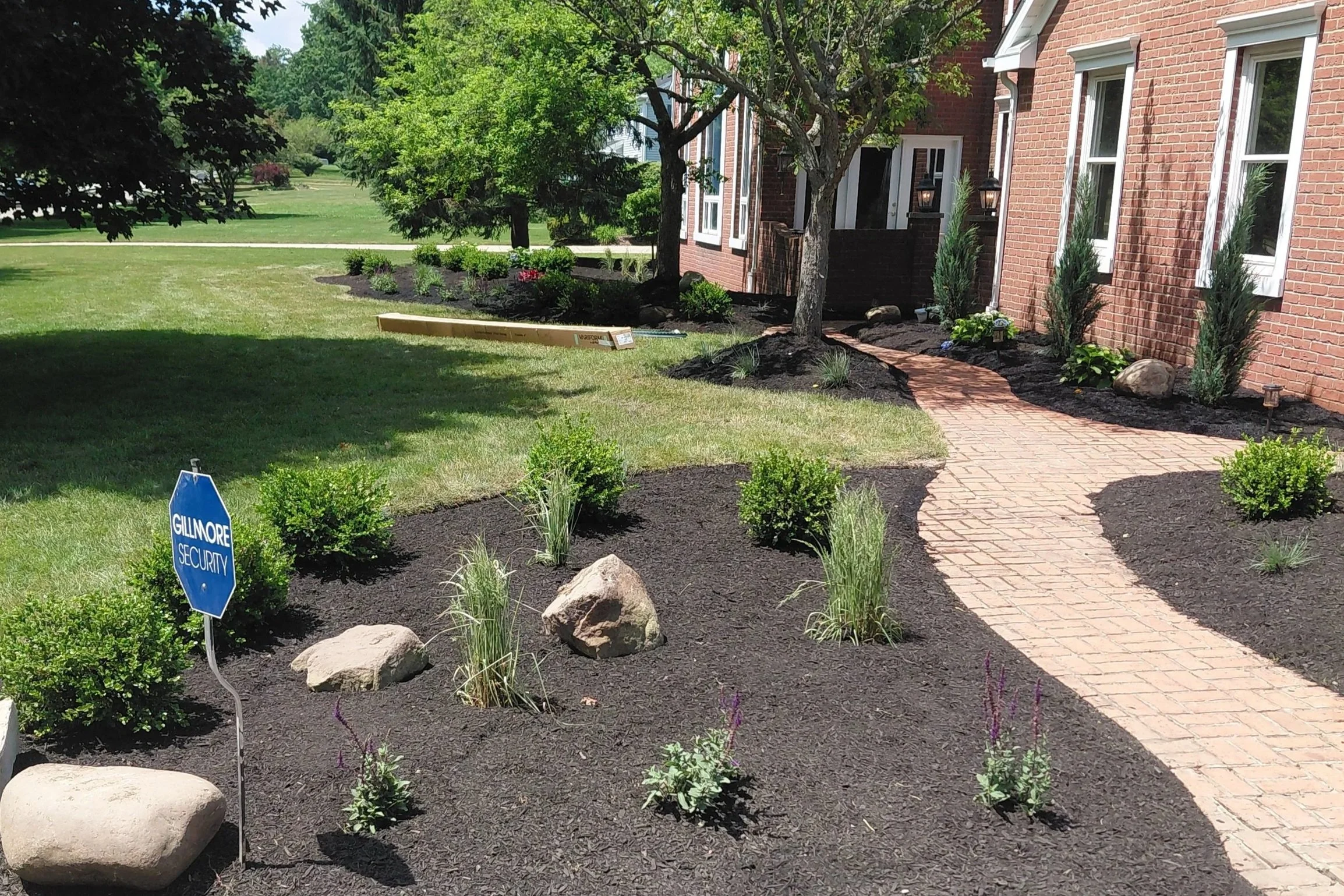 A landscaped front yard featuring a brick house with three white-framed windows and a brick pathway. The yard has freshly planted bushes, trees, and large rocks. There is a shade of a large dark tree on the left, and in the foreground, a blue sign re