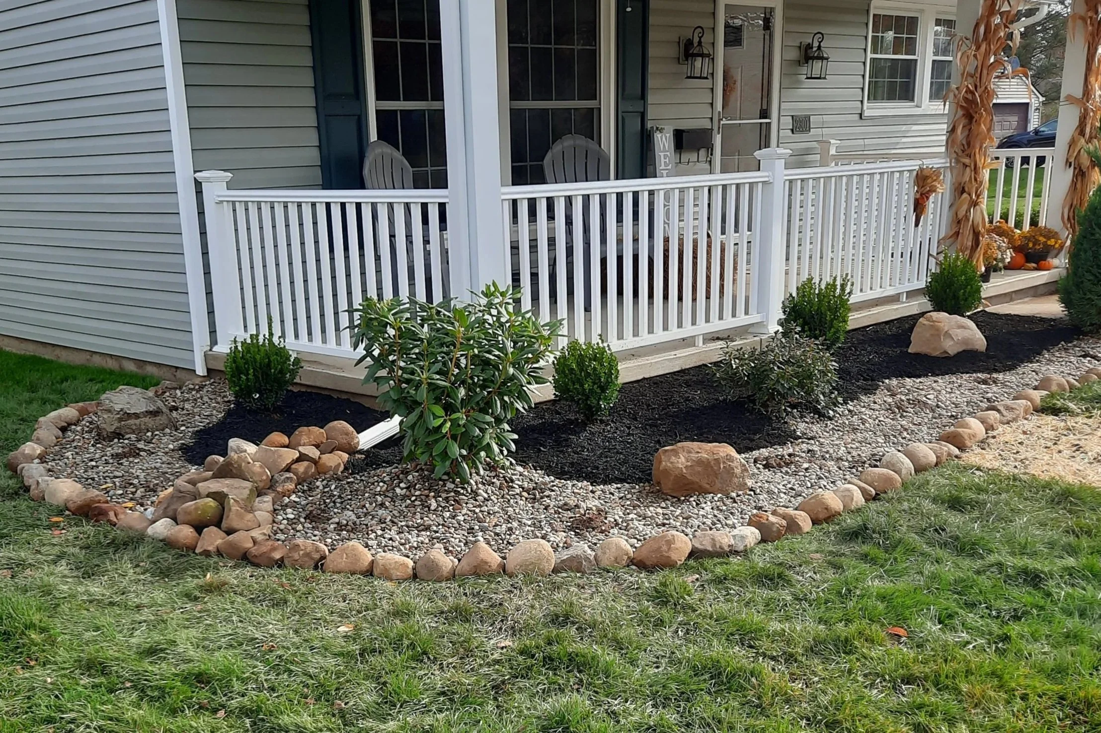 Front porch with white railing and chairs, surrounded by a landscaped garden bed with rocks, small shrubs, and mulch, next to green lawn.