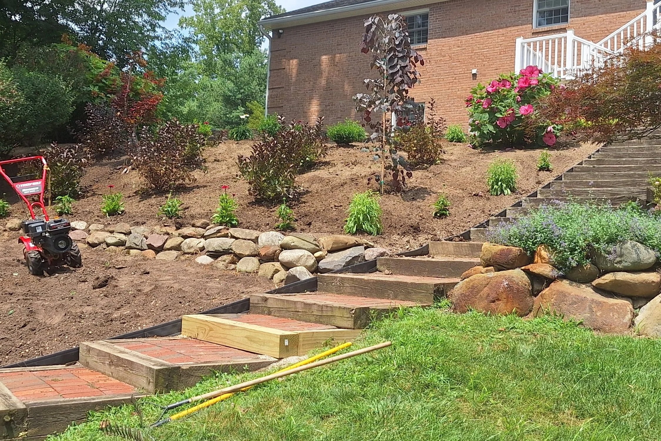 A backyard landscaping scene showing a slope with newly planted bushes, a small tree with dark leaves, colorful flowers, a brick walkway, large rocks, and a brick house in the background with a staircase and white railing.