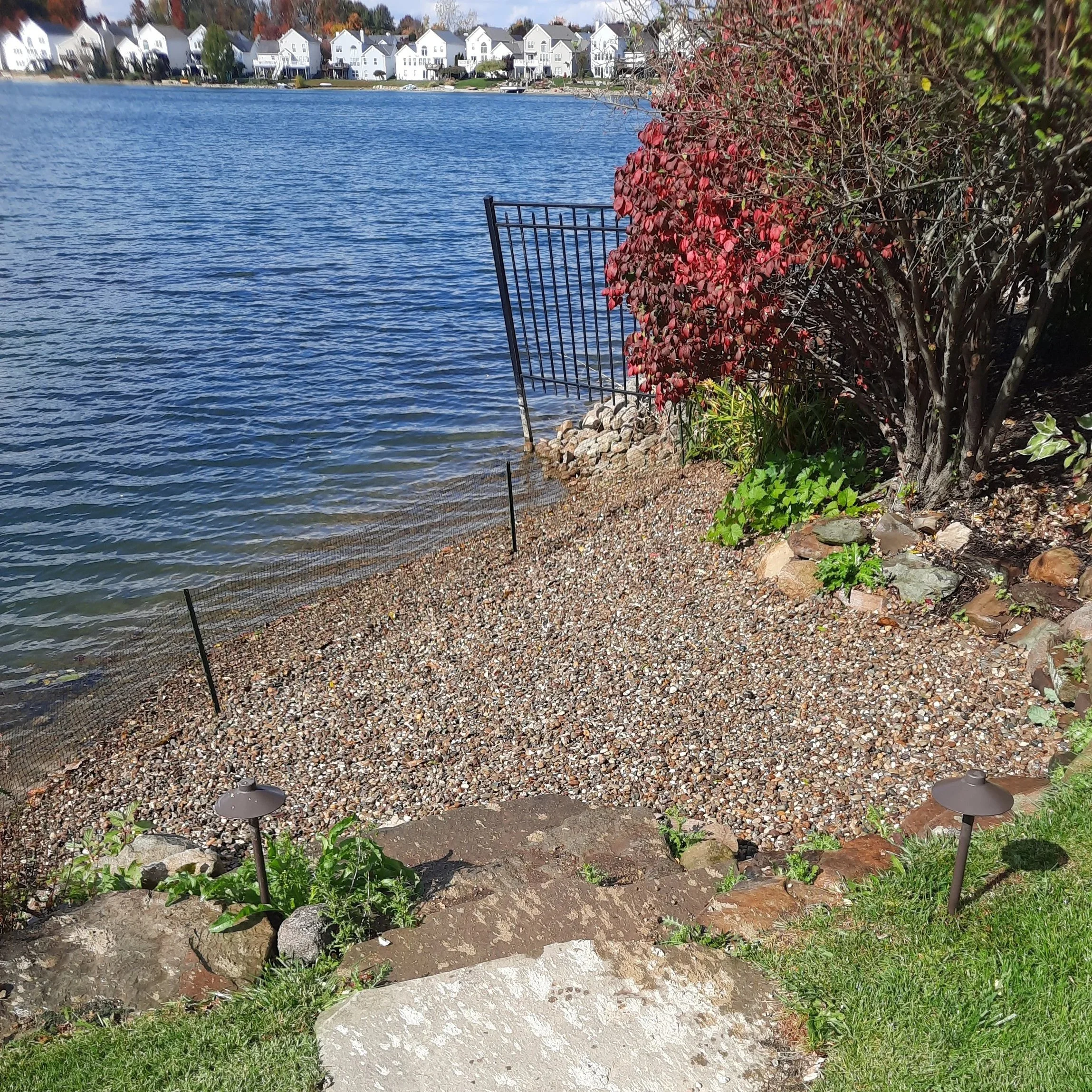 A lakeside backyard with a gravel beach, a metal gate, a small grassy area, and landscape lights, with houses in the background.