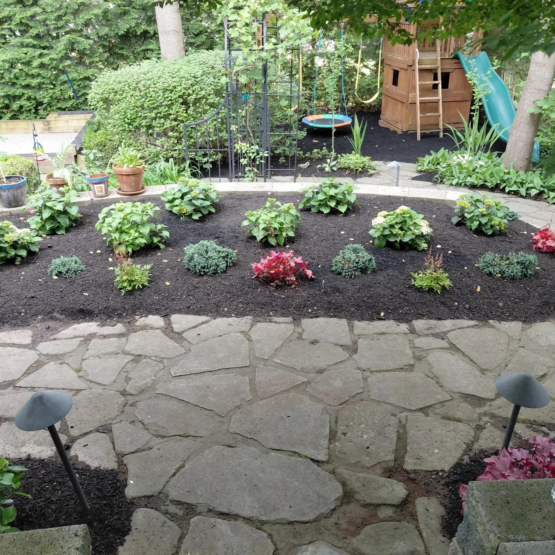 A backyard garden with a stone patio, black mulch, and flower beds with various plants and flowers. In the background, there is a wooden playhouse with a small slide, a ladder, and a treehouse with swings hanging nearby. There are also several potted