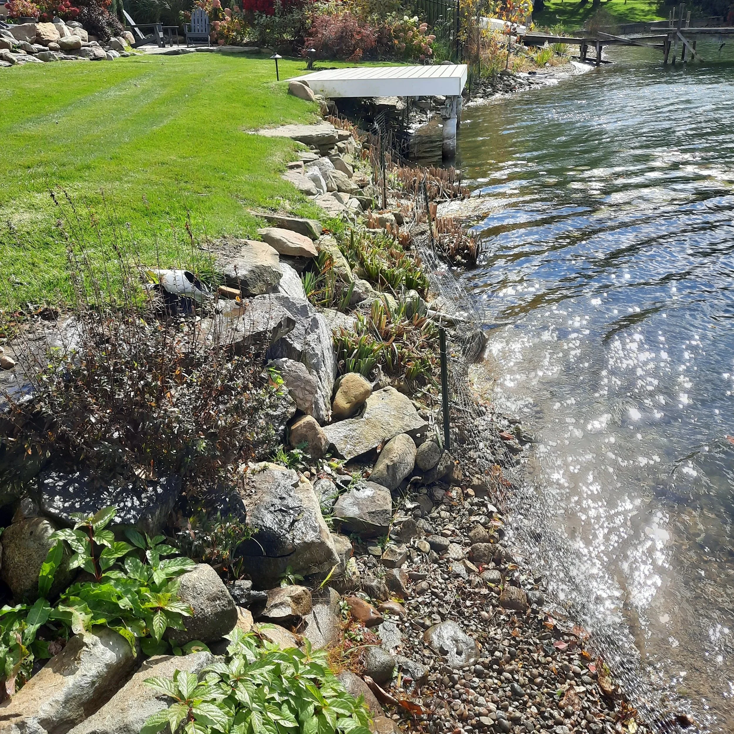 A lakeside yard with a grassy lawn, rocks along the shoreline, and a small white dock extending over the water. There are outdoor chairs and plants near the yard, with trees and flowers in the background.