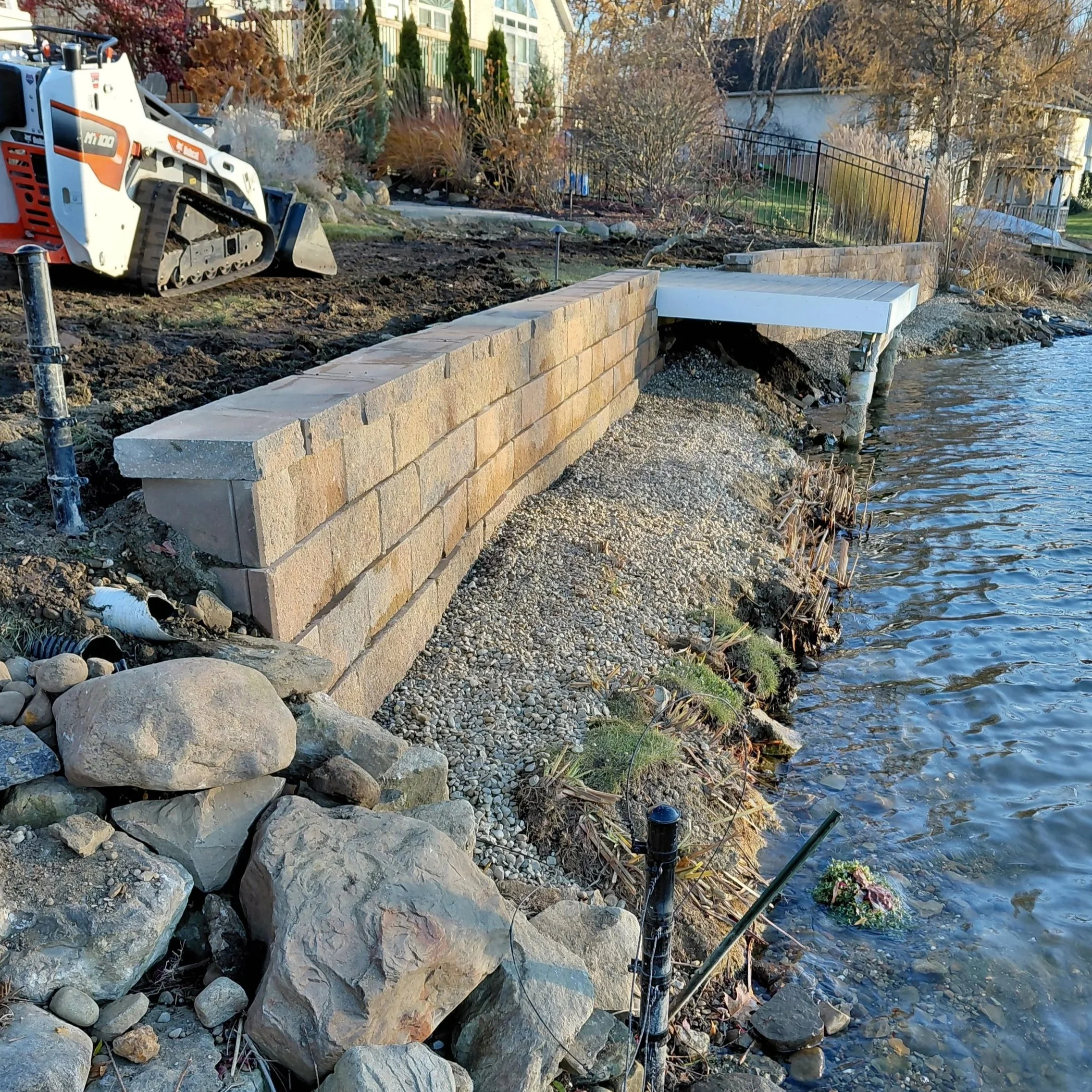 A landscape view of a waterfront area with a newly built stone retaining wall along the shoreline. There is construction equipment, including a small tracked excavator, on the earth behind the wall. The background shows trees, some with autumn foliag