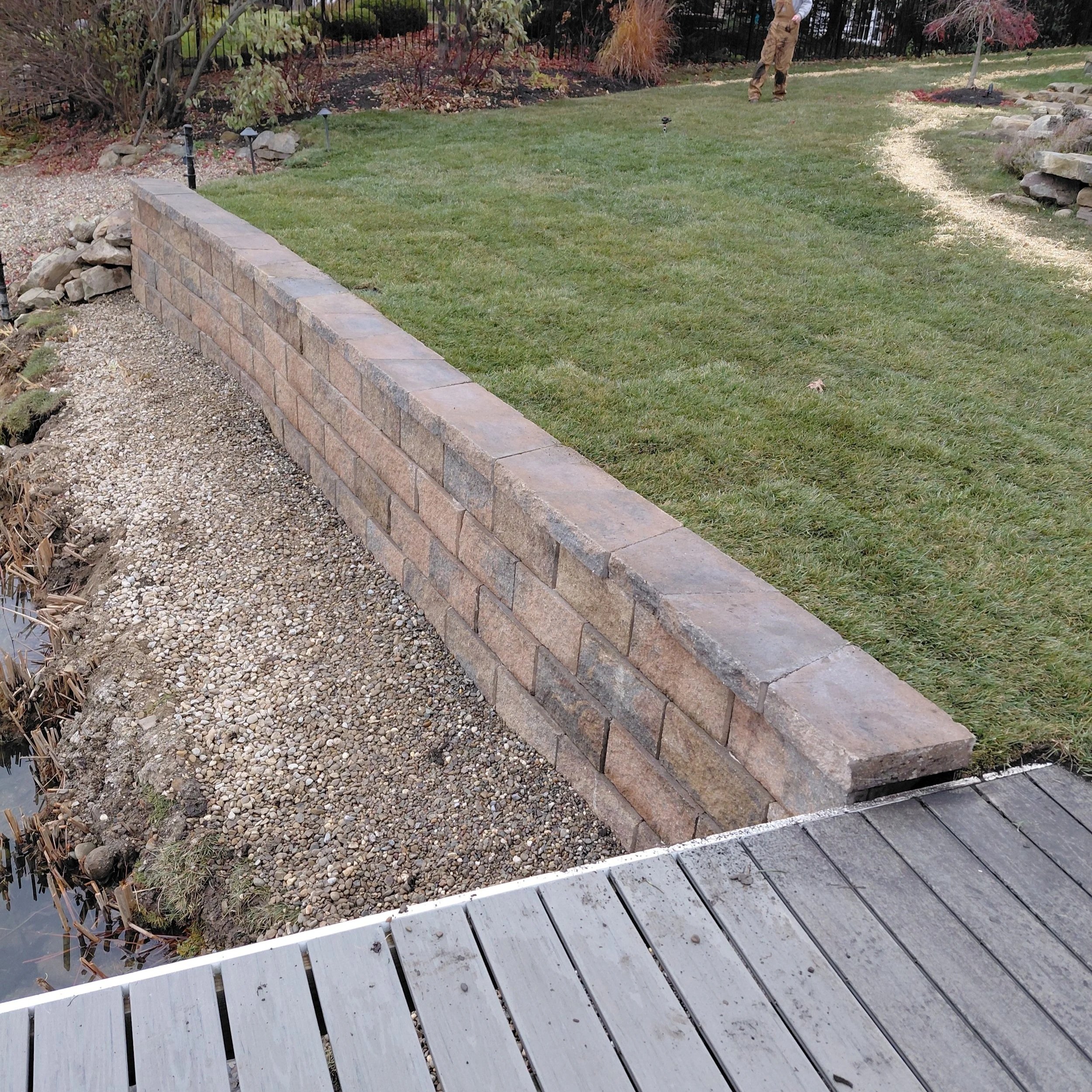 A backyard garden with a short brick wall, a grassy lawn, a curved path, and a person standing in the background. The garden has plants, bushes, and small landscape lights.