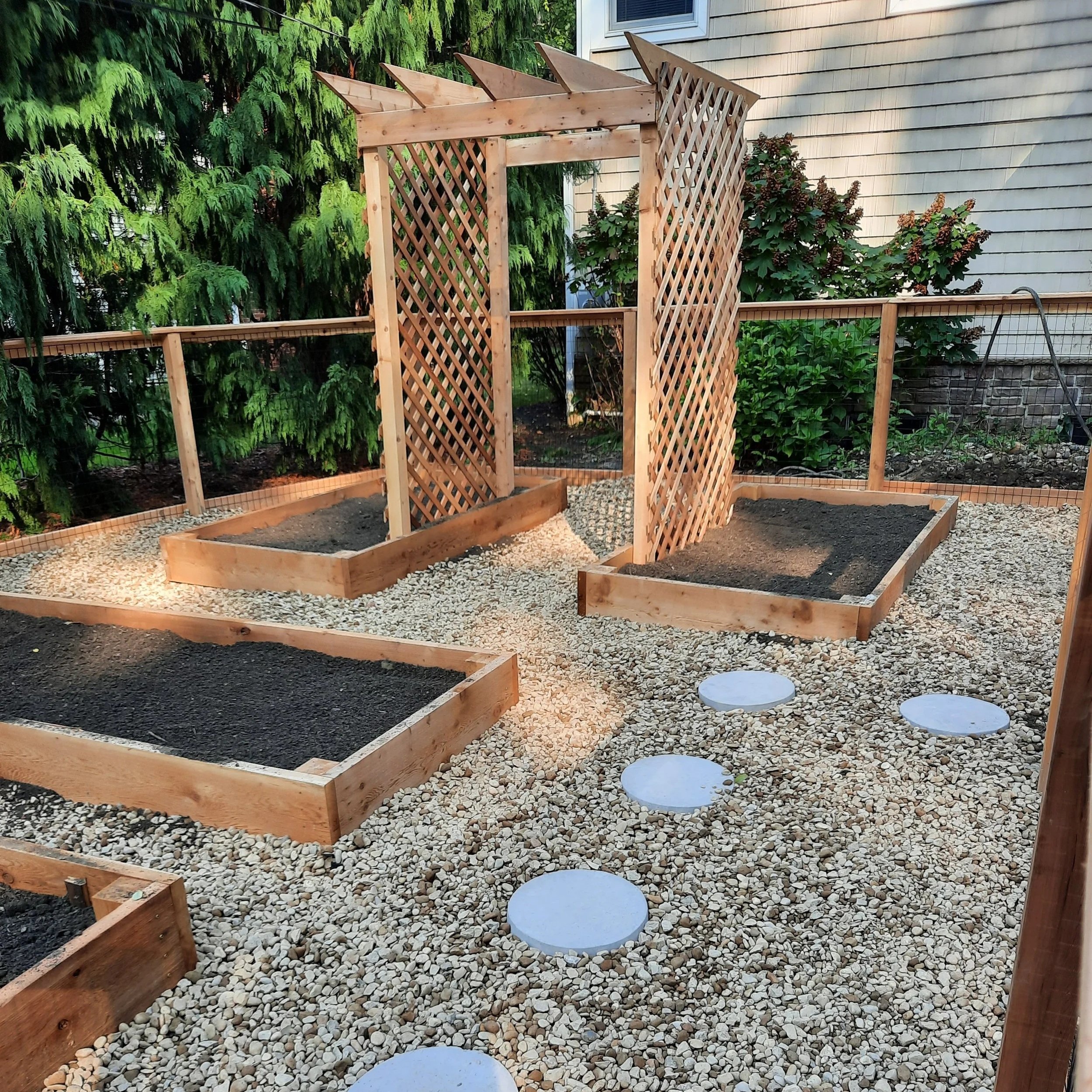 A small backyard garden with a wooden trellis structure for climbing plants, surrounded by shrubbery and greenery, with some planting beds and stepping stones.