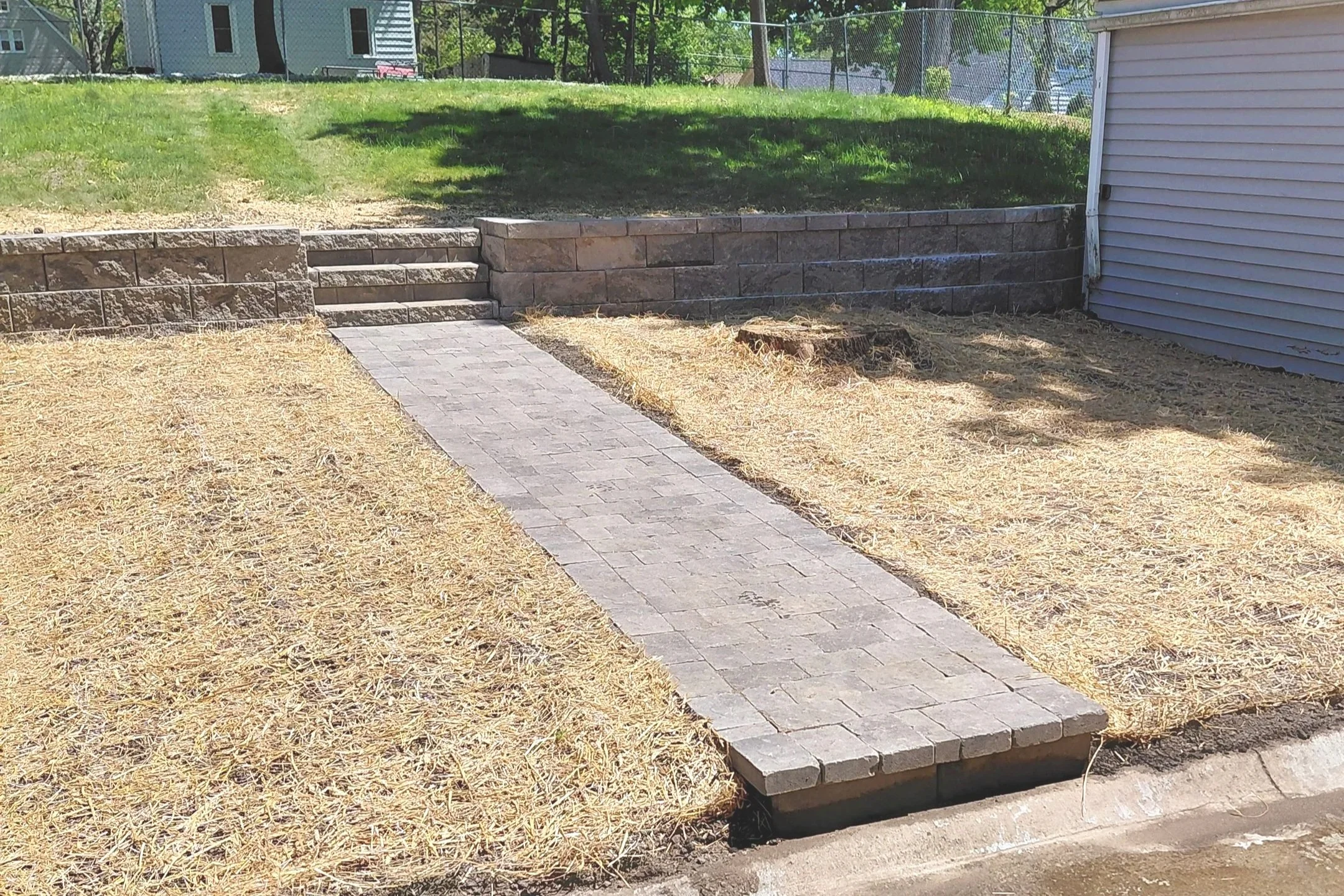 A backyard with a small brick walkway leading to a tiered stone retaining wall, a grassy area, a tree, a house, a fence, and a tree stump.