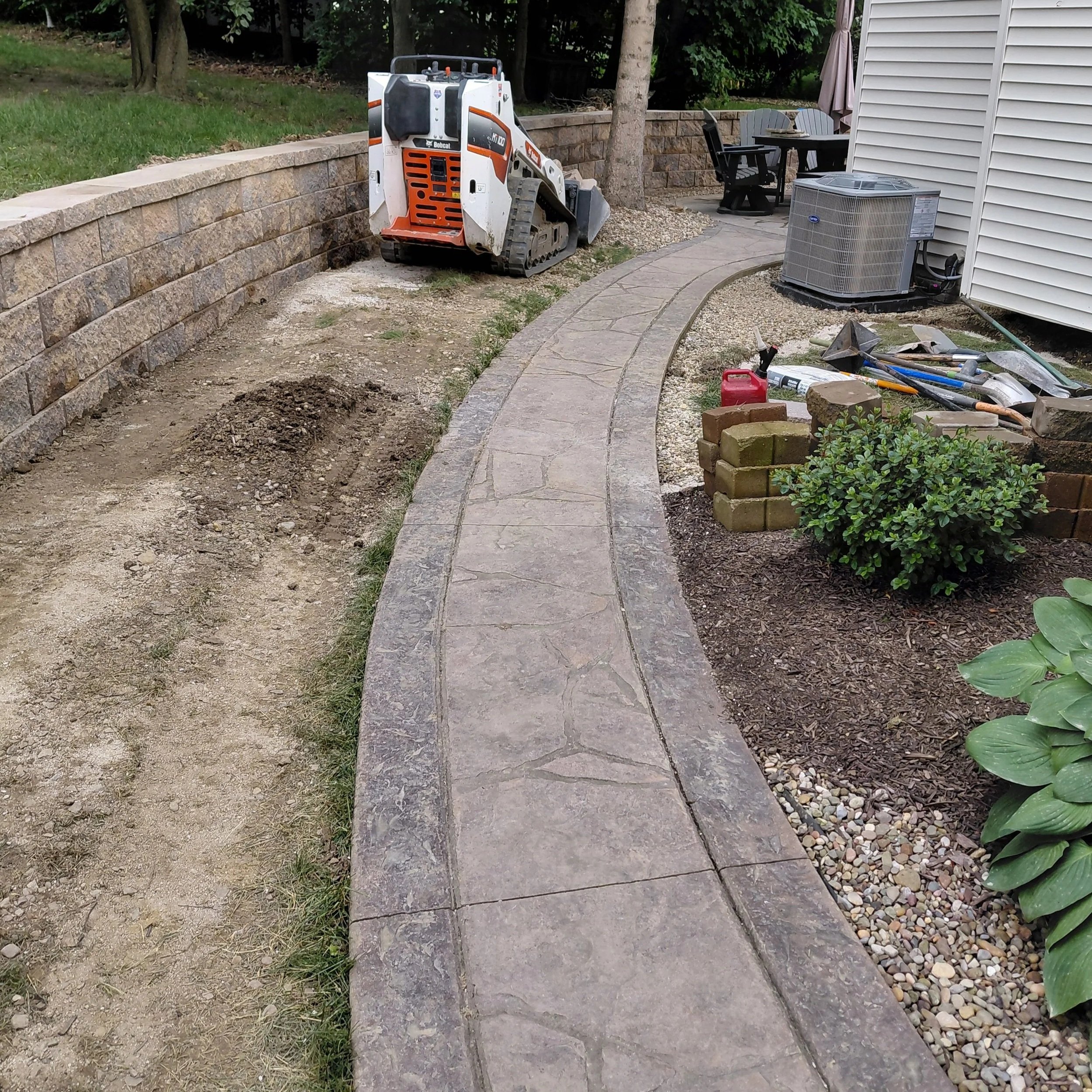 A small construction or landscaping equipment with tracks, located near a curved brick walkway in a backyard. There is a patio area with outdoor chairs, an air conditioning unit, and scattered tools and materials.