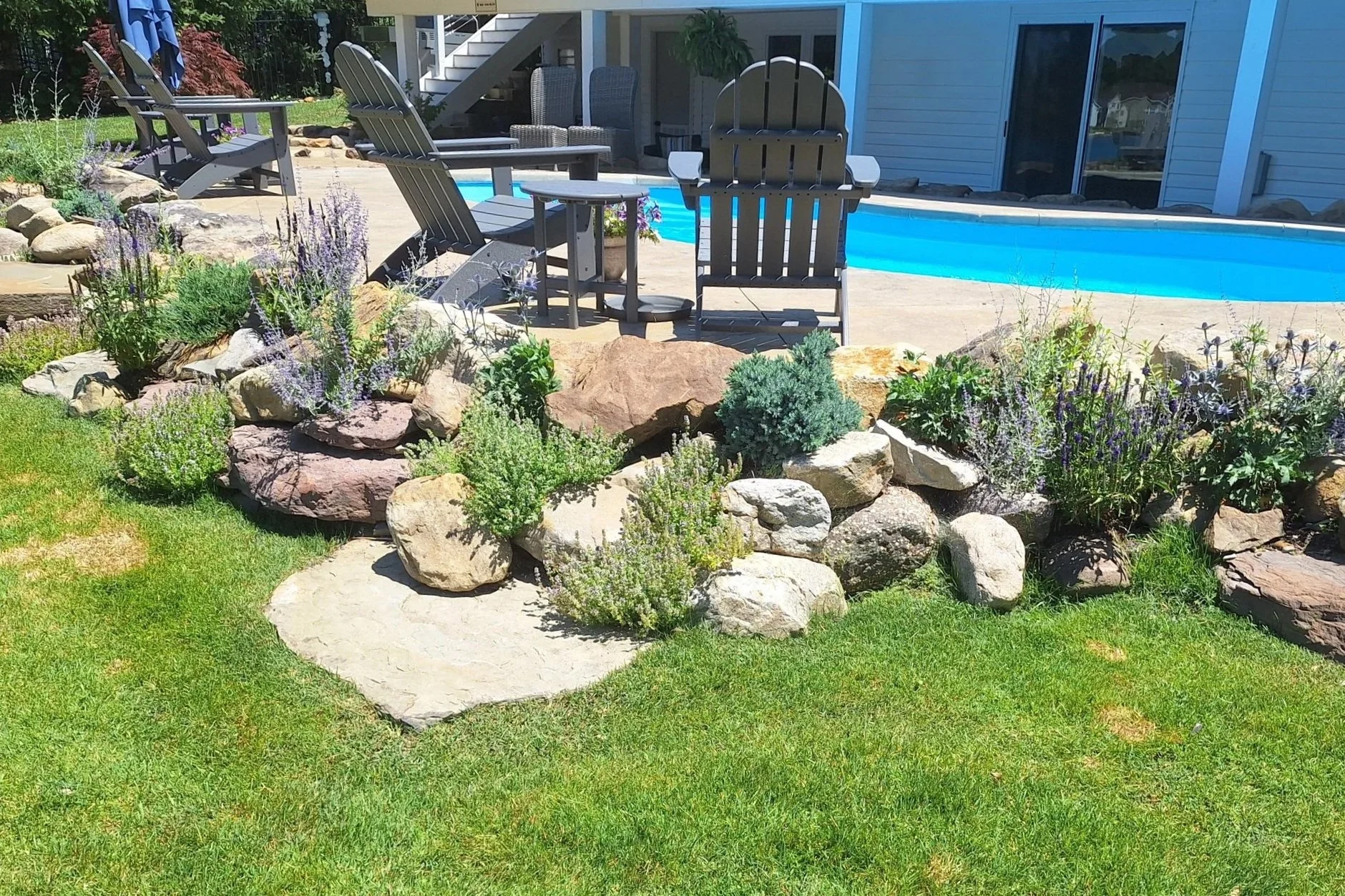 A backyard with a landscaped garden featuring rocks, flowering plants, and a grassy area in the foreground. In the background, there's a swimming pool, outdoor seating, and a house with steps leading to an upper level.