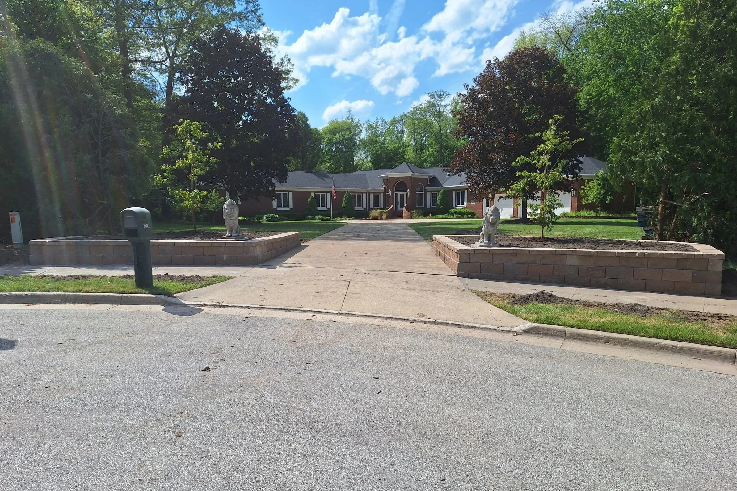 A suburban house with a brick facade and a gray roof, situated behind a landscaped yard with two lion statues on brick platforms, trees, and a driveway leading to a garage on the right.