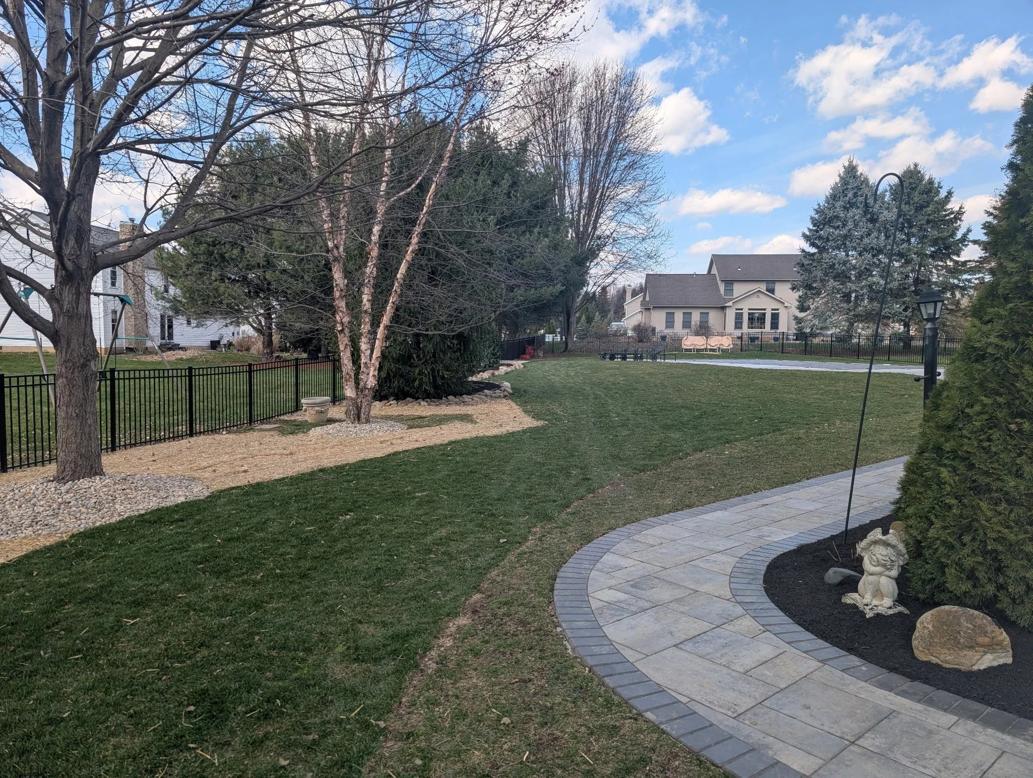 Backyard with lawn, trees, a curved stone patio, a decorative statue of a dog, and a black metal fence. In the background are houses, trees, and a cloudy sky.