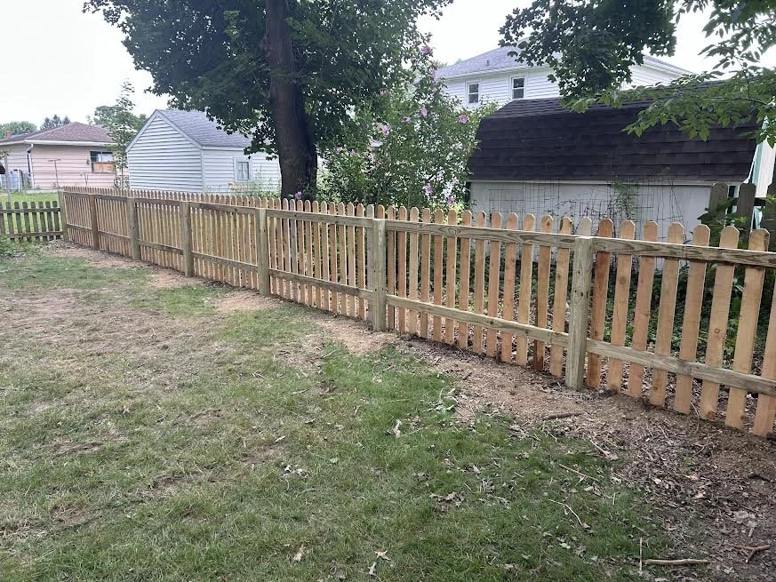 A freshly installed wooden picket fence runs along a grassy backyard with a large tree and neighboring houses in the background.