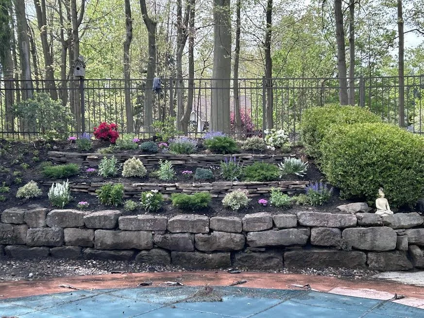 Yard with a multi-tiered stone garden bed filled with various shrubs and flowering plants, bordered by a black metal fence, with trees in the background and a small Buddha statue on the right side.