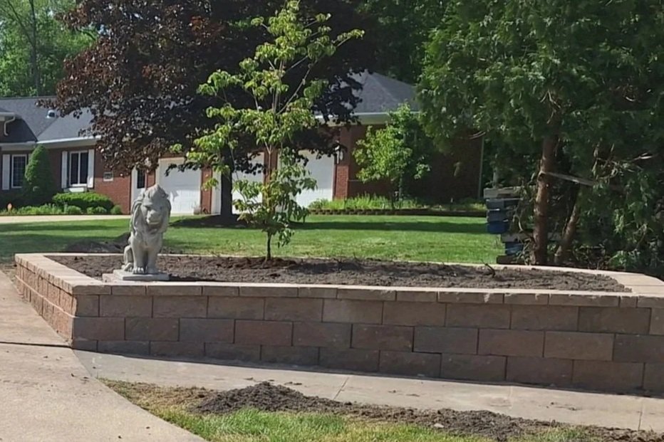 A small tree is planted in a raised brick garden bed, with a dog statue at its base, in a residential neighborhood with houses and trees in the background.