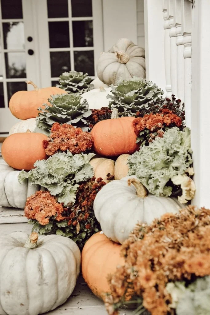 A collection of pumpkins and ornamental cabbage plants arranged outdoors near a white porch, featuring white, orange, and gray pumpkins among green and orange foliage.