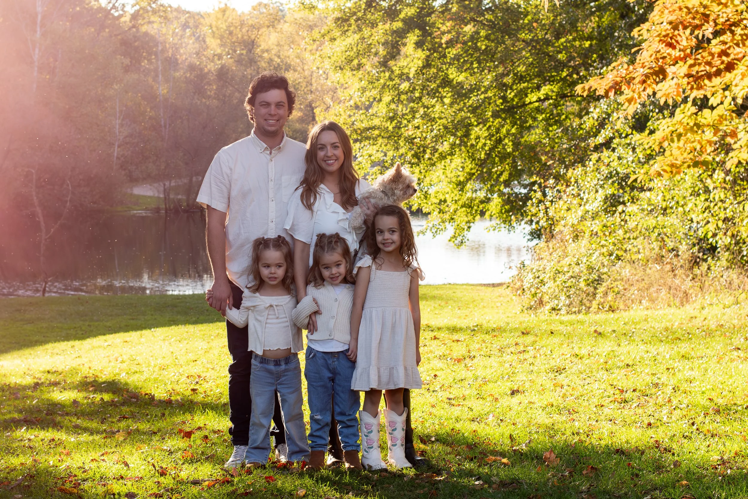 Family of six posing outdoors near a lake with autumn trees in the background. The family includes two adult men, one adult woman, and three young girls. One girl is holding a small dog.