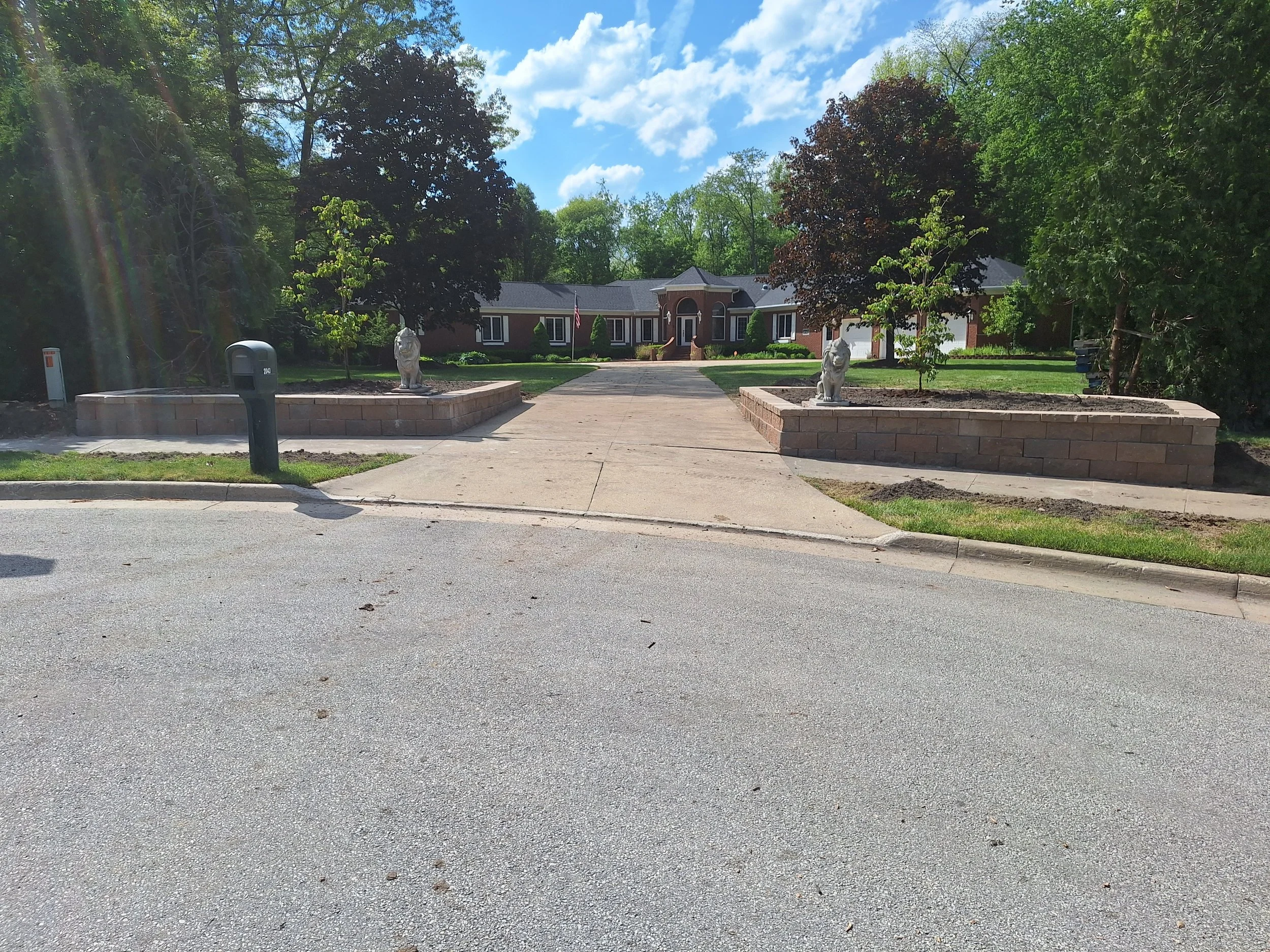 A large brick house with a driveway leading to the entrance, flanked by two raised flower beds with statues of lions, trees, and a blue sky with clouds.