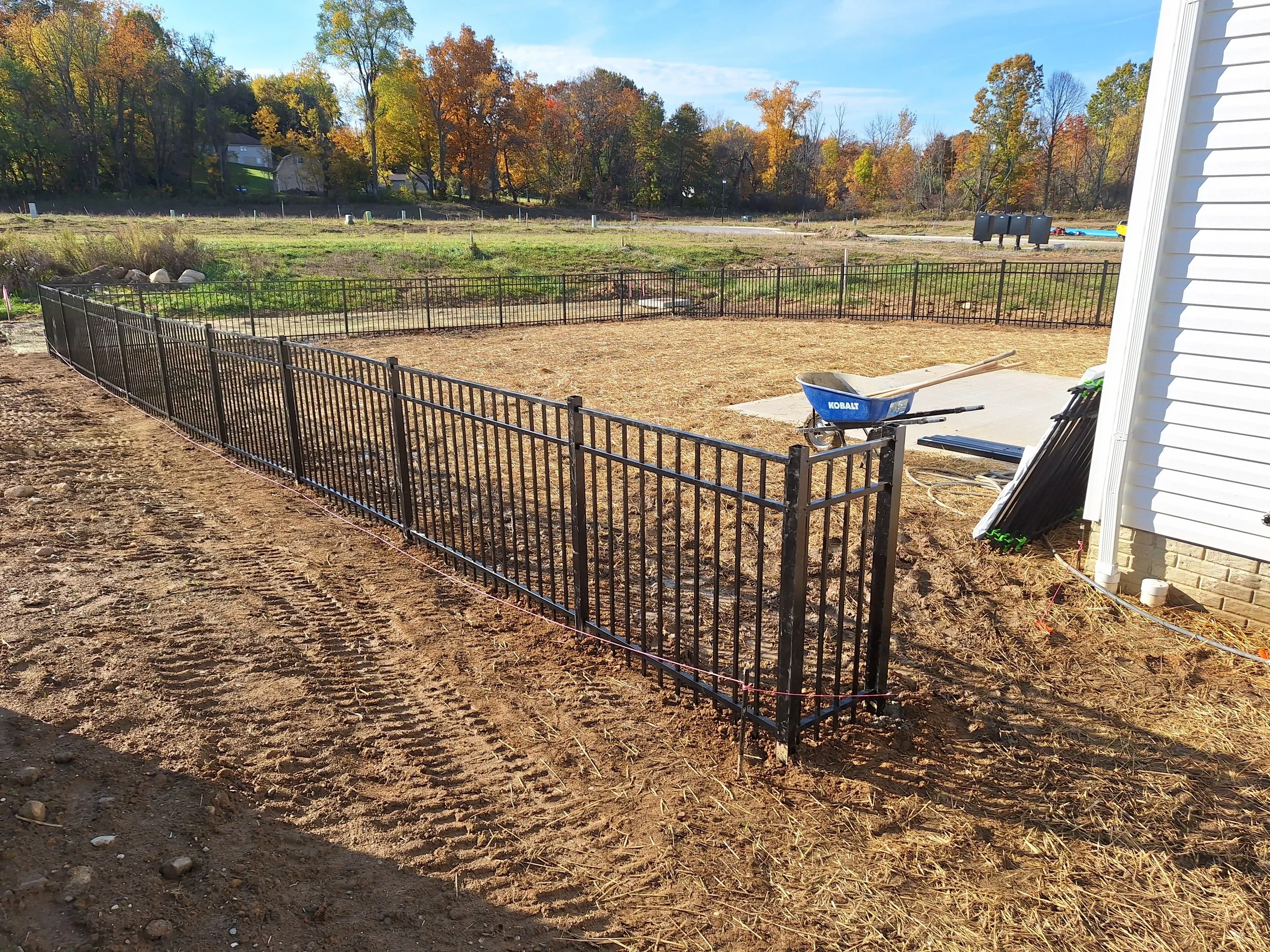 A backyard with a newly installed black metal fence surrounding a section of soil. Construction equipment, including a blue wheelbarrow, is present. In the background, there are trees with autumn foliage and some residential houses.