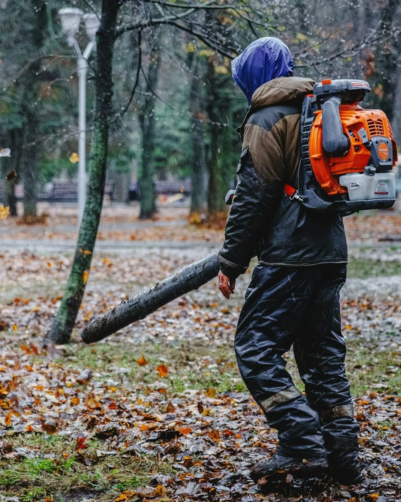 A person wearing rain gear, including a hooded jacket and waterproof pants, is carrying a leaf blower and holding a fallen tree branch in a park with wet leaves and trees.