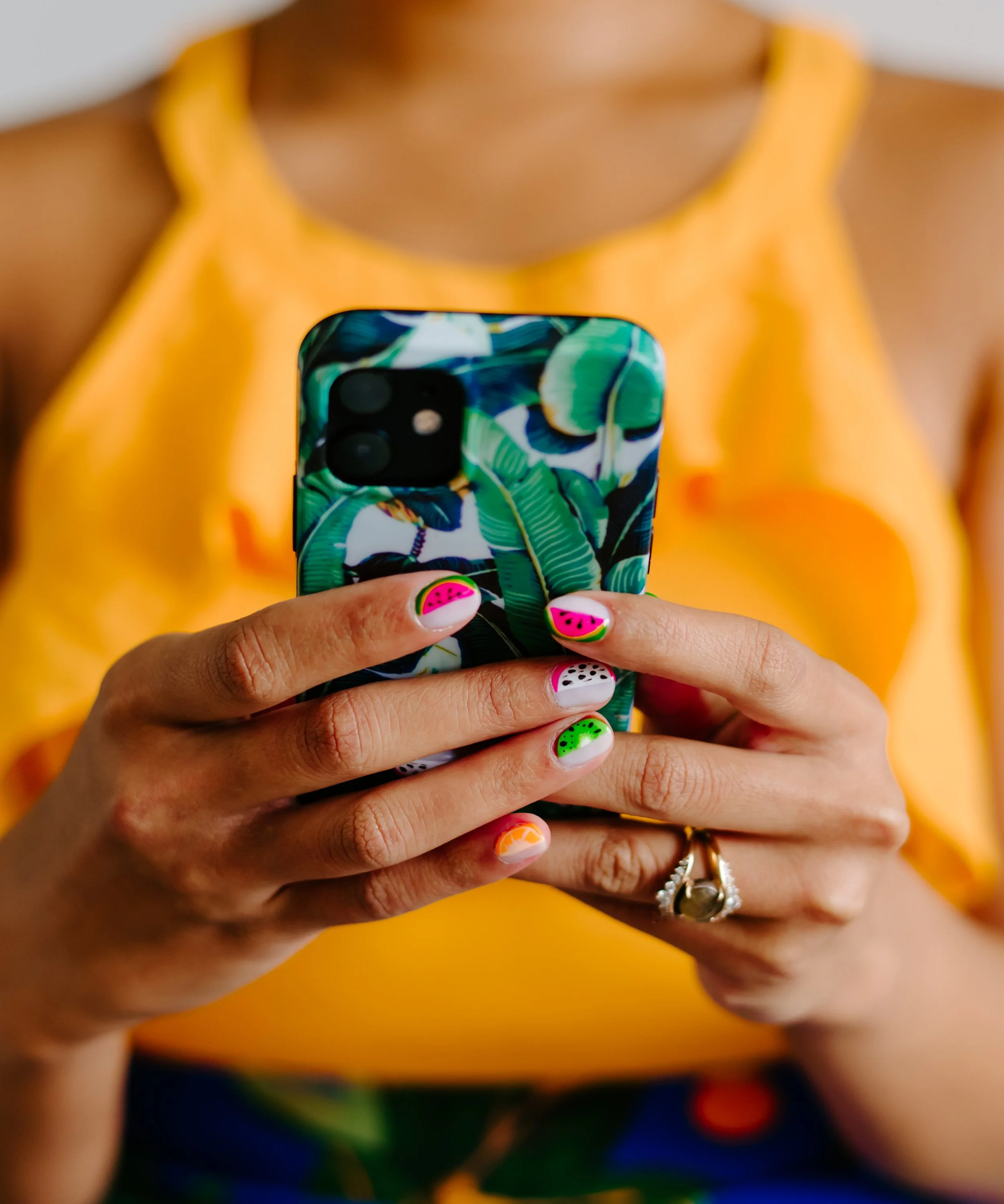 A person holding a smartphone with a tropical leaf pattern case. Finger nails painted with watermelon and fruit designs, rings visible on fingers. Person wearing a yellow top, background blurred.