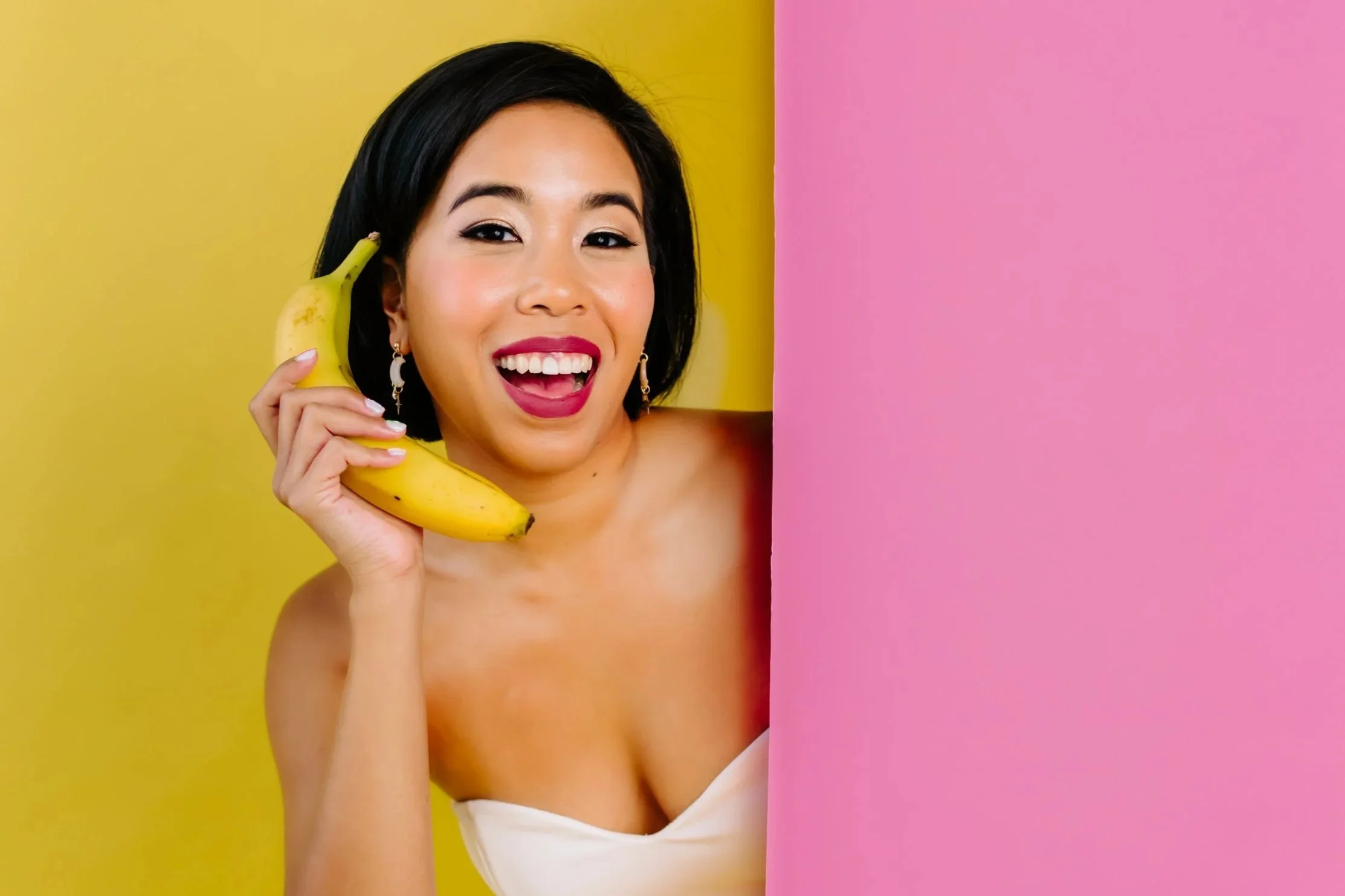 A woman with black hair and earrings holding a banana to her ear, smiling at the camera, with a yellow background on the left and a pink background on the right.