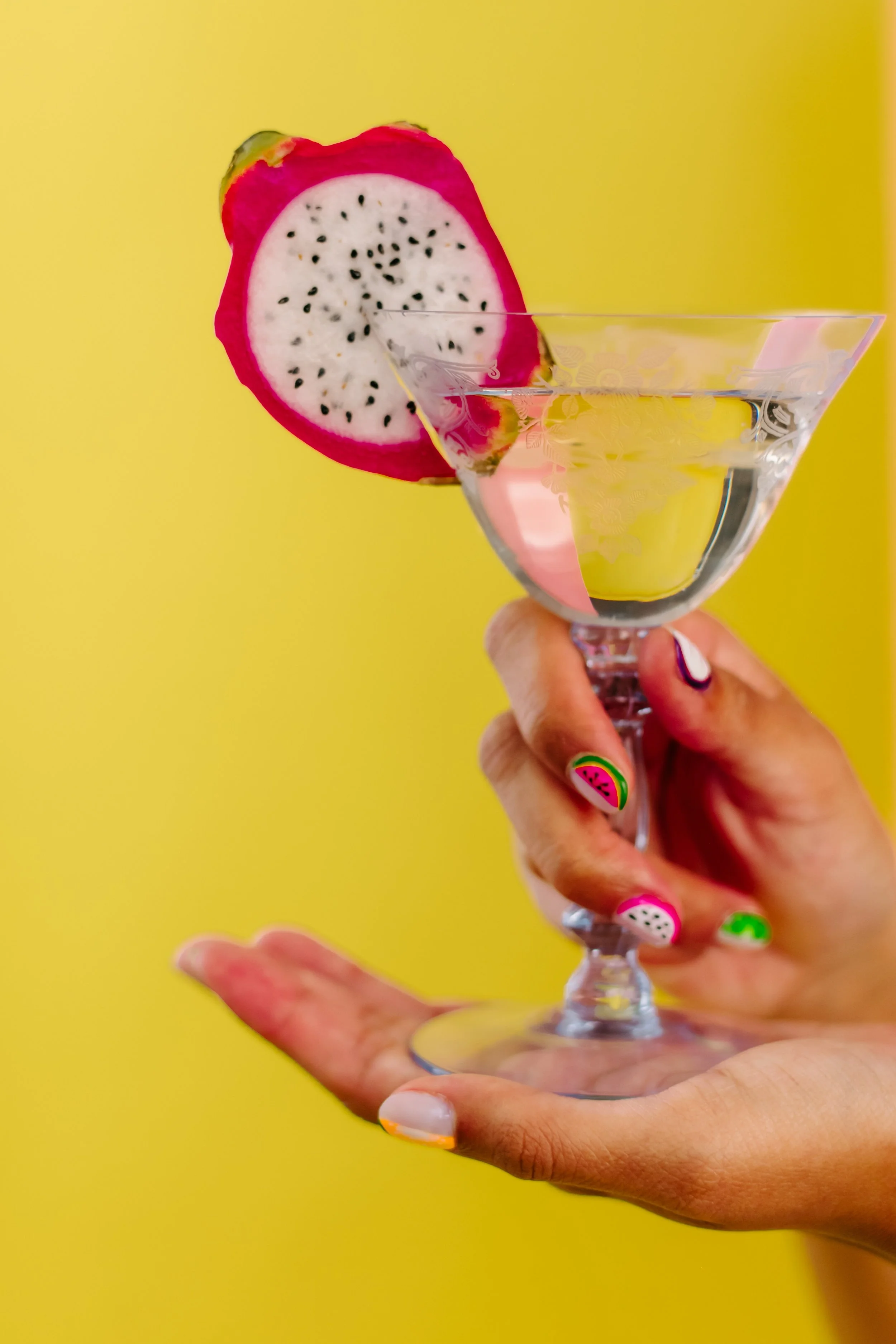 A hand holding a glass with a slice of dragon fruit on a yellow background.